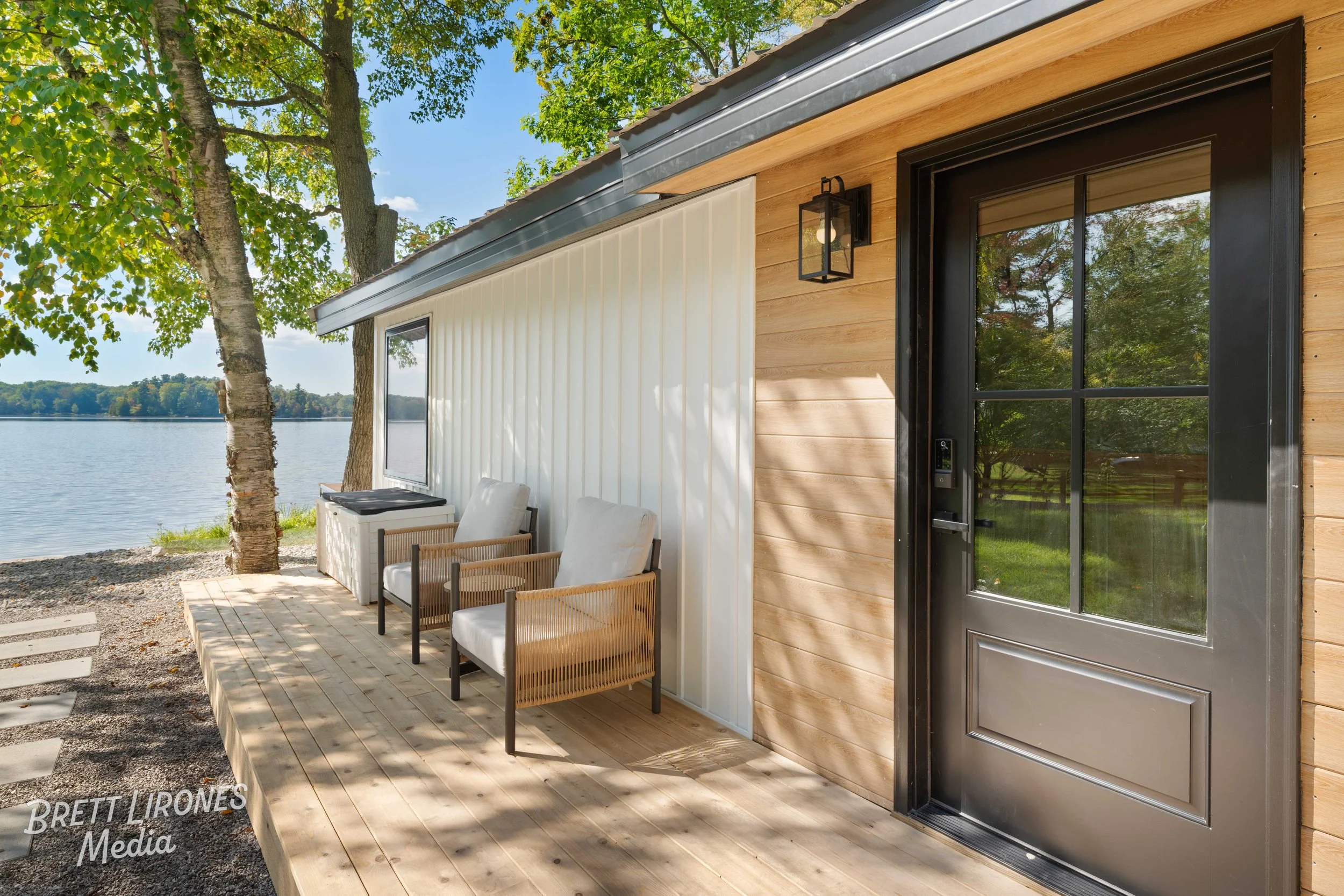 Front porch of a modern house overlooking a lake, with two chairs, an outdoor table, and a large window reflecting trees.