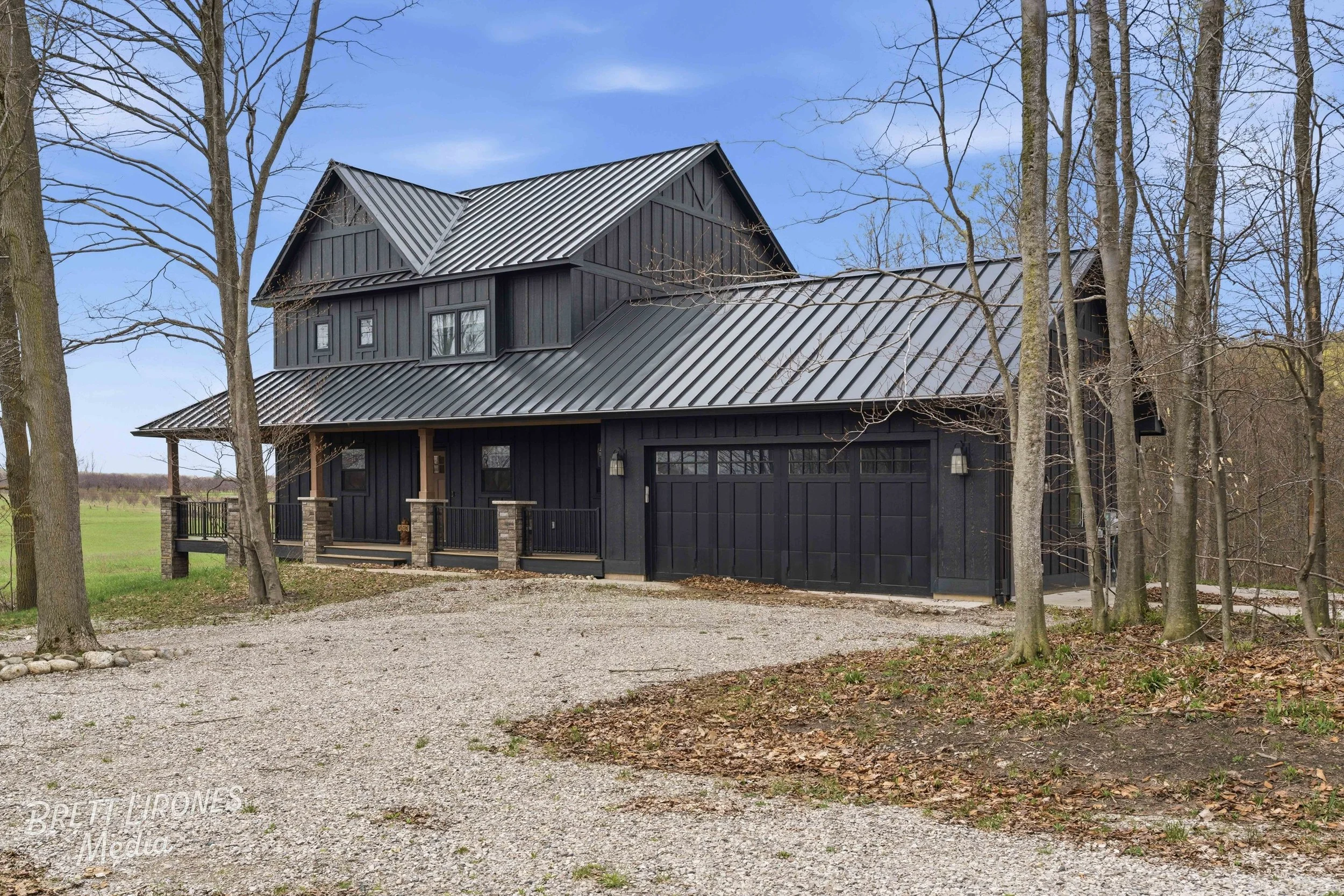 A modern black house with a metal roof, surrounded by leafless trees, with a gravel driveway in the foreground and a grassy field in the background.