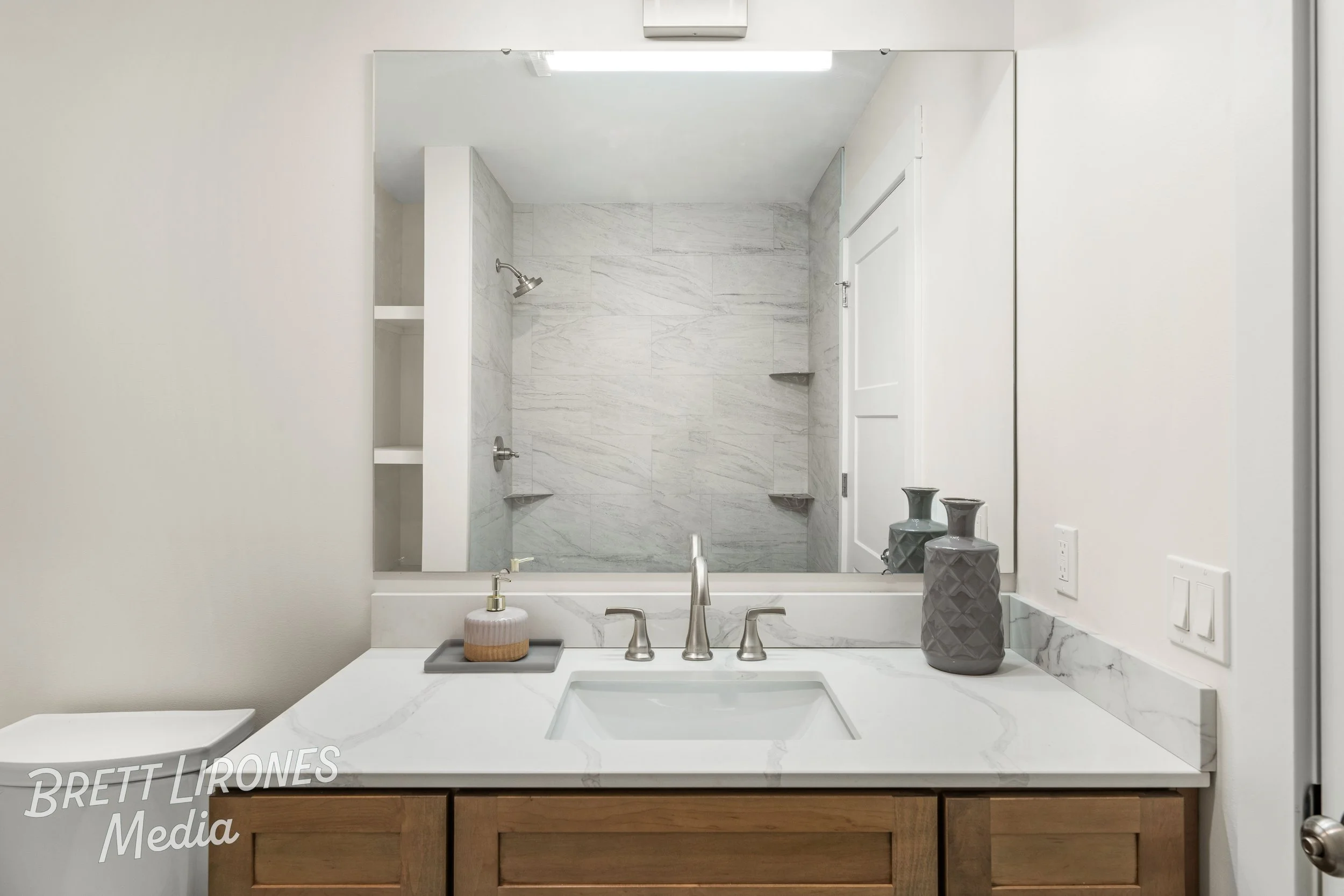 Bathroom vanity with a white marble countertop, a gray vase and soap dispenser, and a large mirror reflecting a walk-in shower with marble walls and corner shelves.