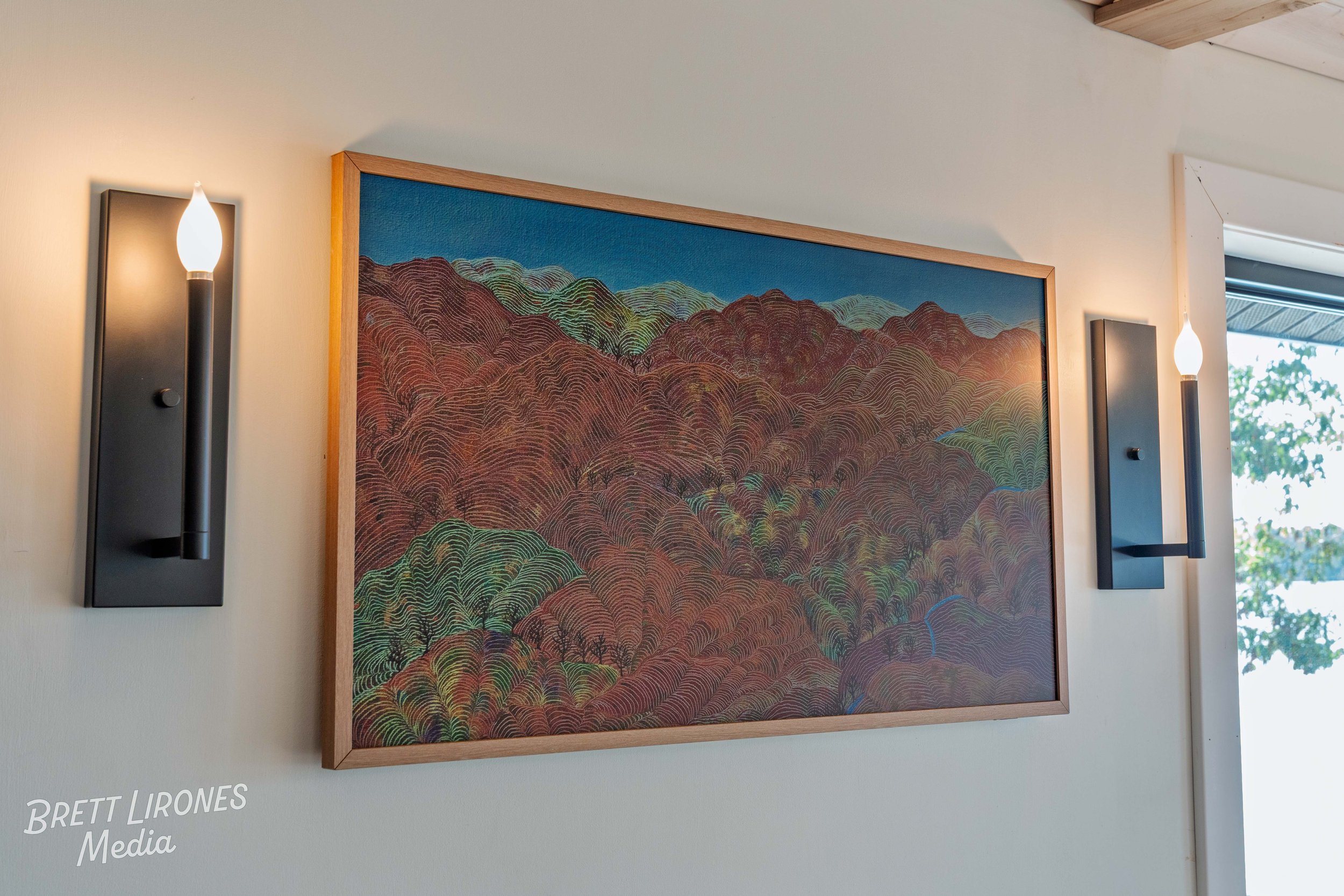 Interior wall with two modern black wall sconces and a framed colorful landscape painting of mountains and trees, next to a window showing greenery outside.