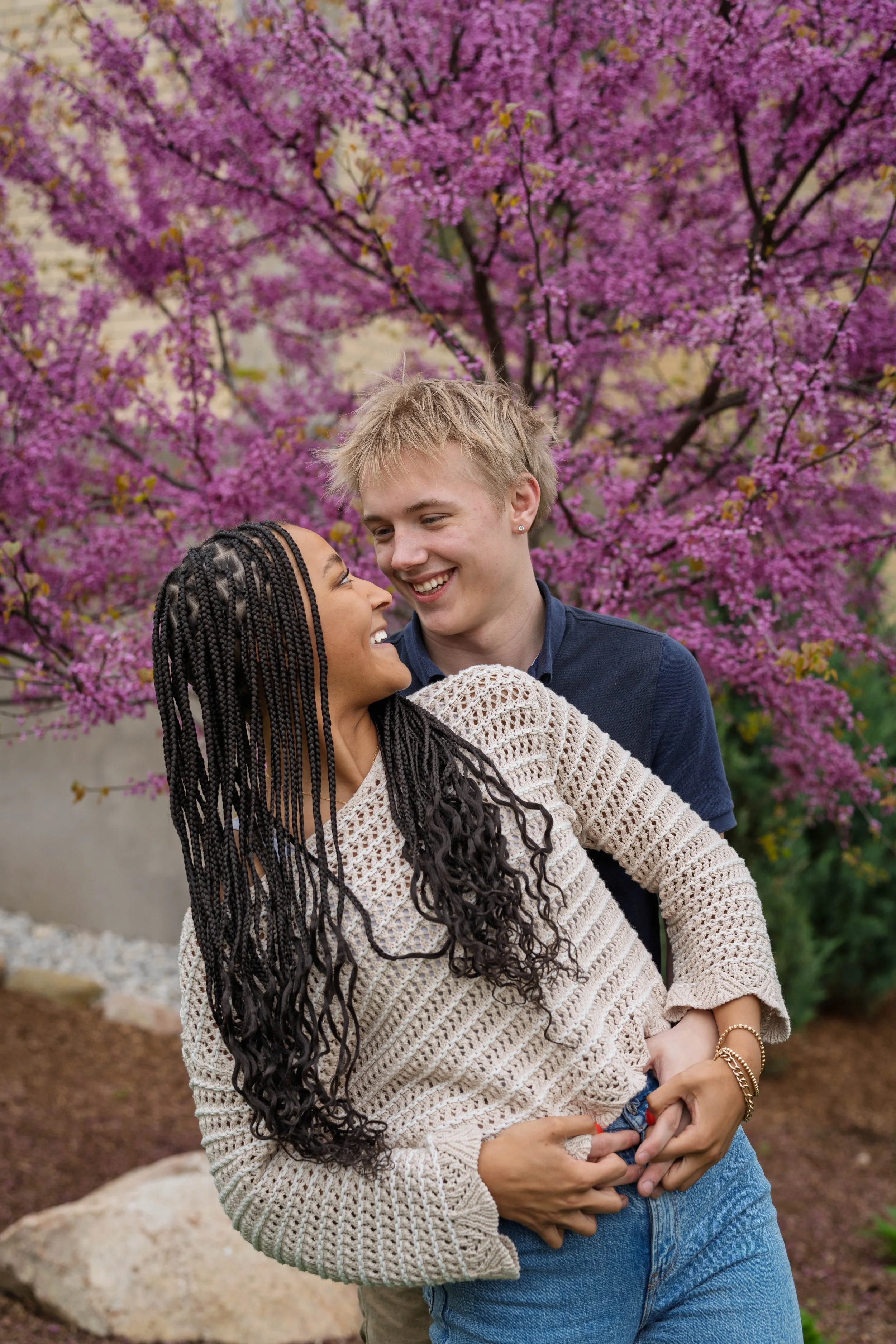 A smiling couple embracing in front of pink flowering trees in a park.