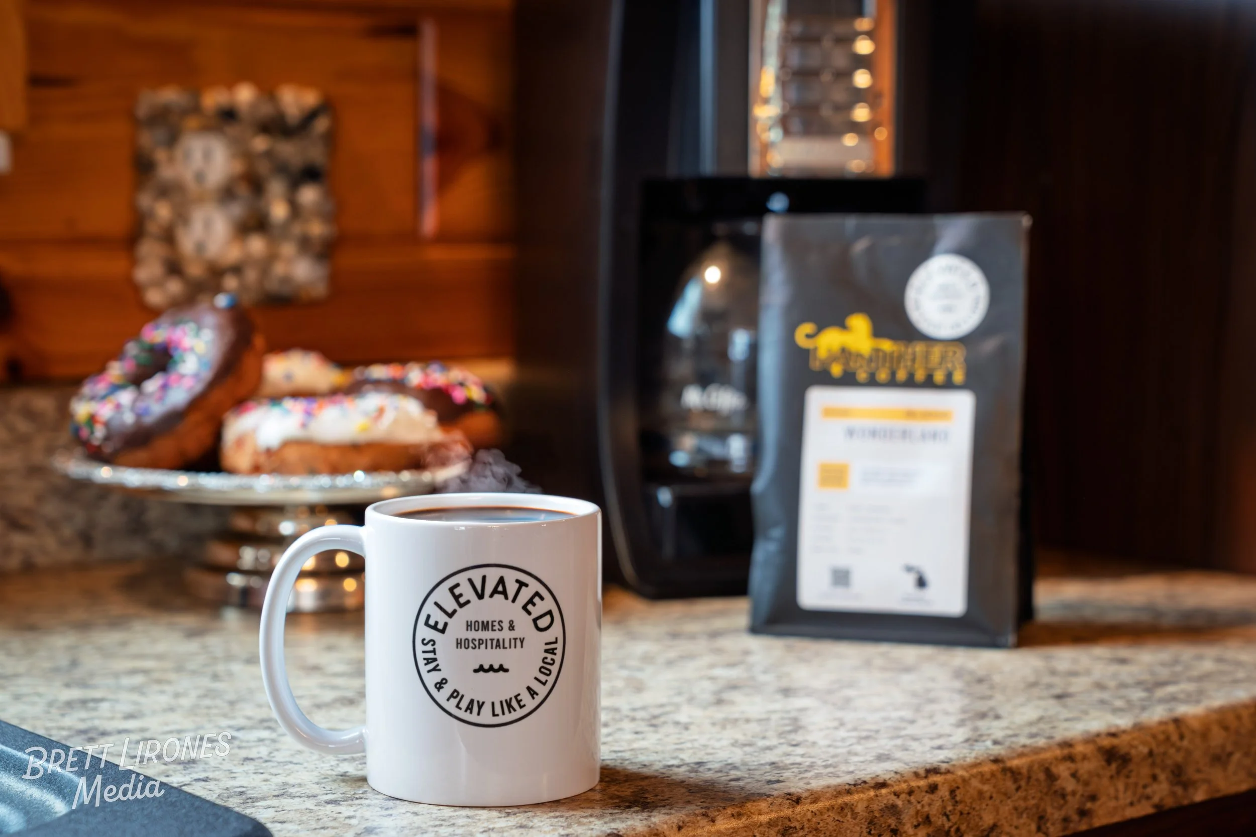 A white coffee mug with a black circular logo saying "Elevated Homes & Hospitality & Play Like a Local" on a kitchen counter, with a plate of assorted donuts with colorful sprinkles and a bag of Killer Coffee in the background, next to a coffee machi