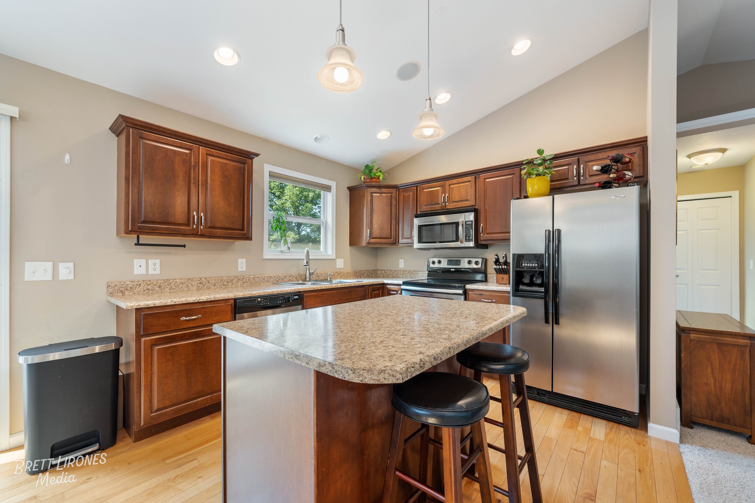 Kitchen with brown wooden cabinets, granite countertops, stainless steel refrigerator, microwave, oven, and a small island with two black stools, under pendant lights, with a window showing greenery outside.