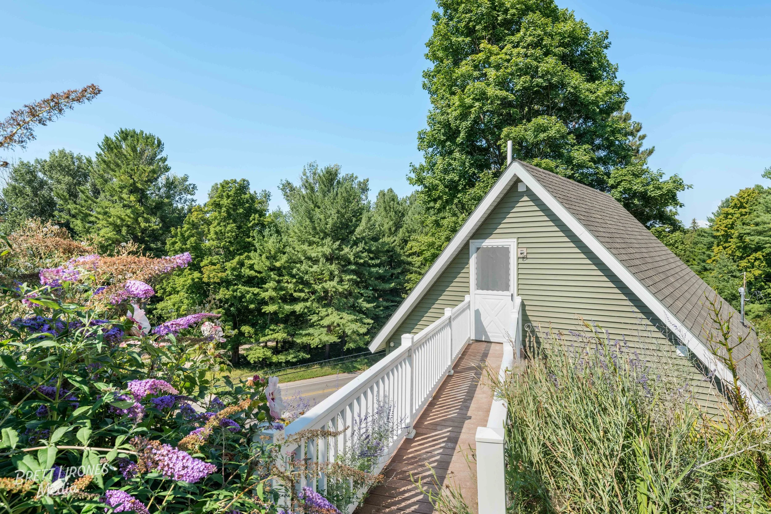 A house with a light green exterior and a steep gray roof, with a white door at the top of a small outdoor staircase, surrounded by greenery and plants, under a clear blue sky.