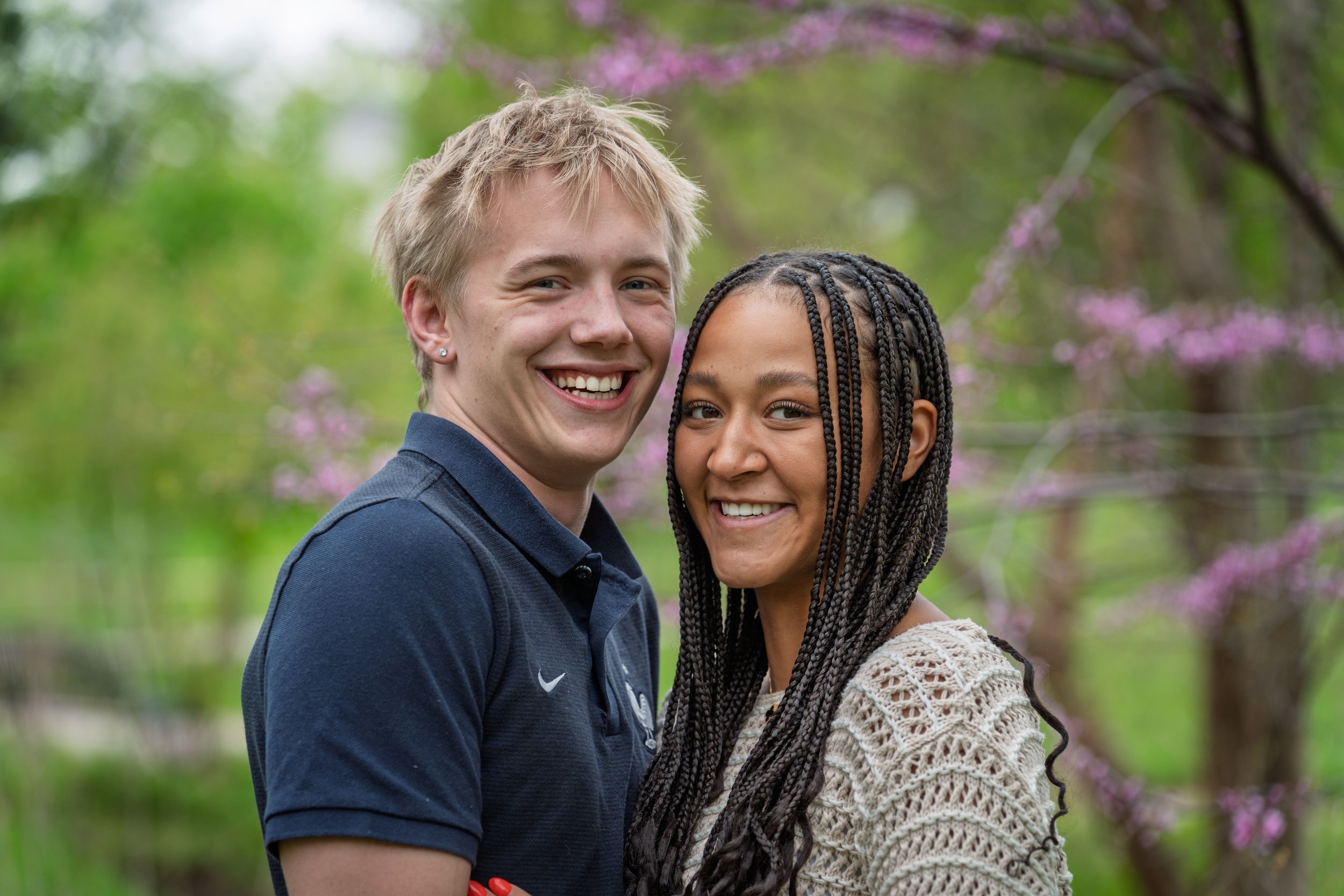 A smiling young man and woman standing close together outdoors with pink flowering trees and green foliage in the background.