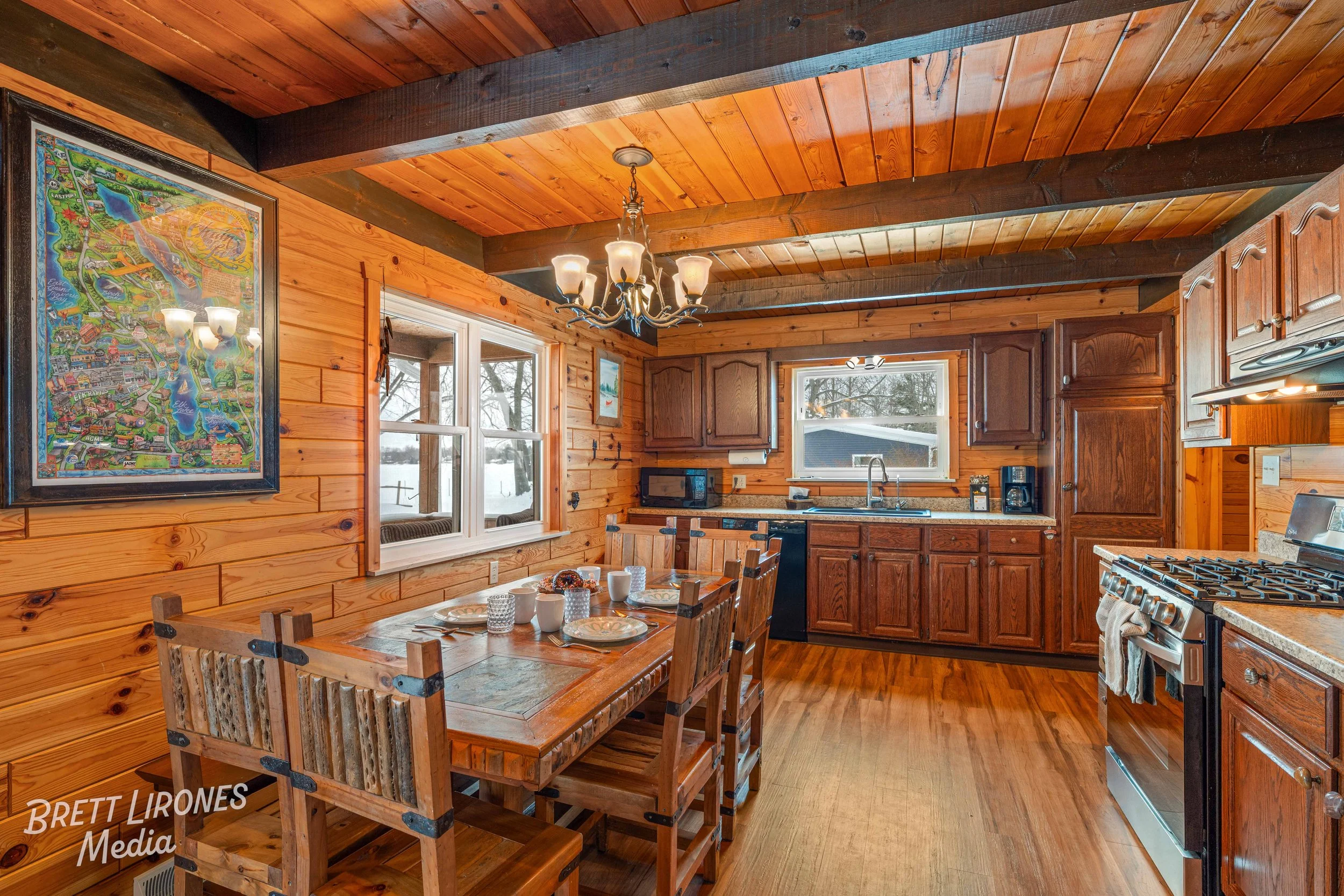 Rustic wooden kitchen with dining table, six chairs, windows showing snowy outdoor scene, hanging chandelier, and kitchen appliances.