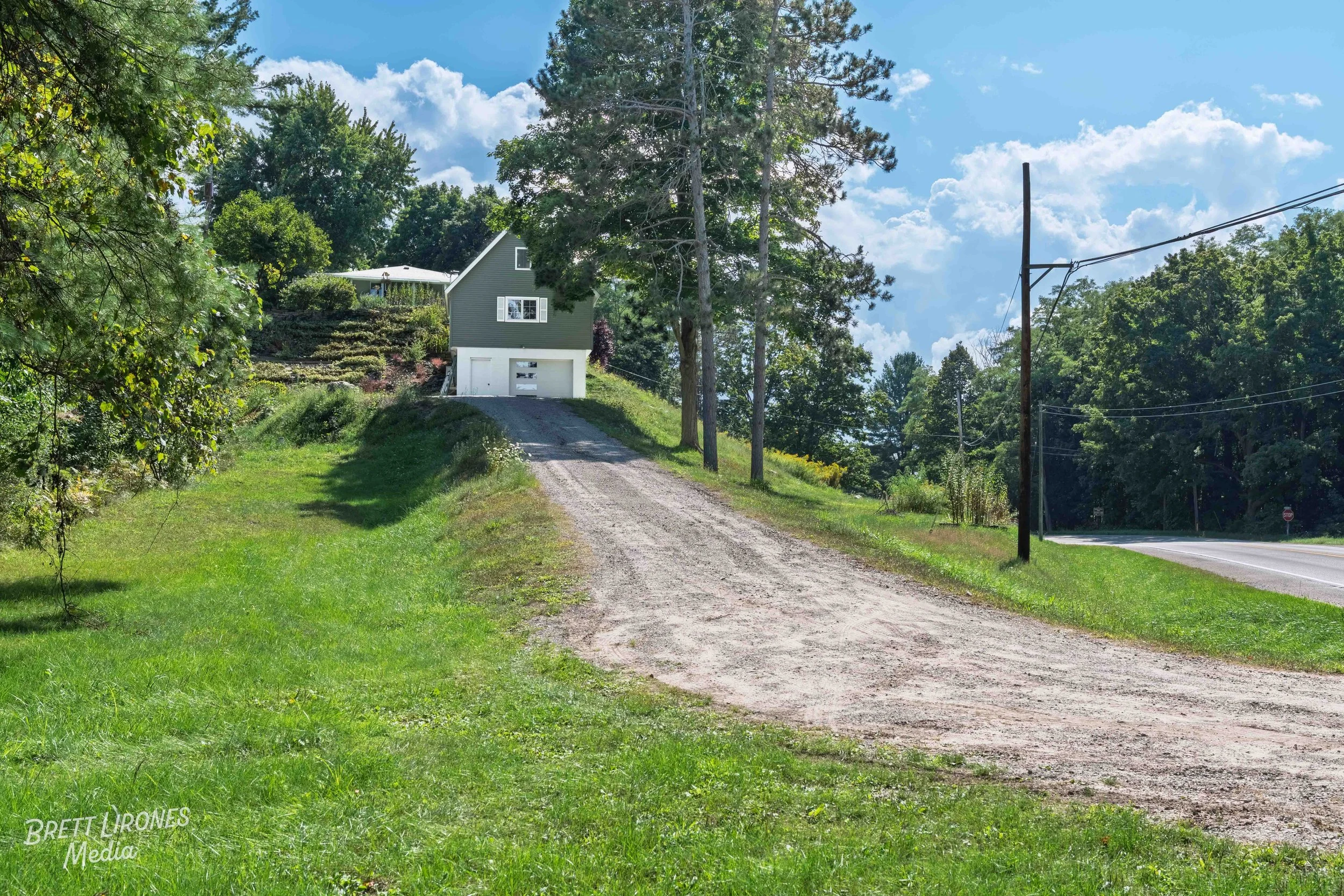 A house on a hill with a dirt driveway leading to a garage. The house is green with white trim, surrounded by tall trees, with a partly cloudy sky overhead.