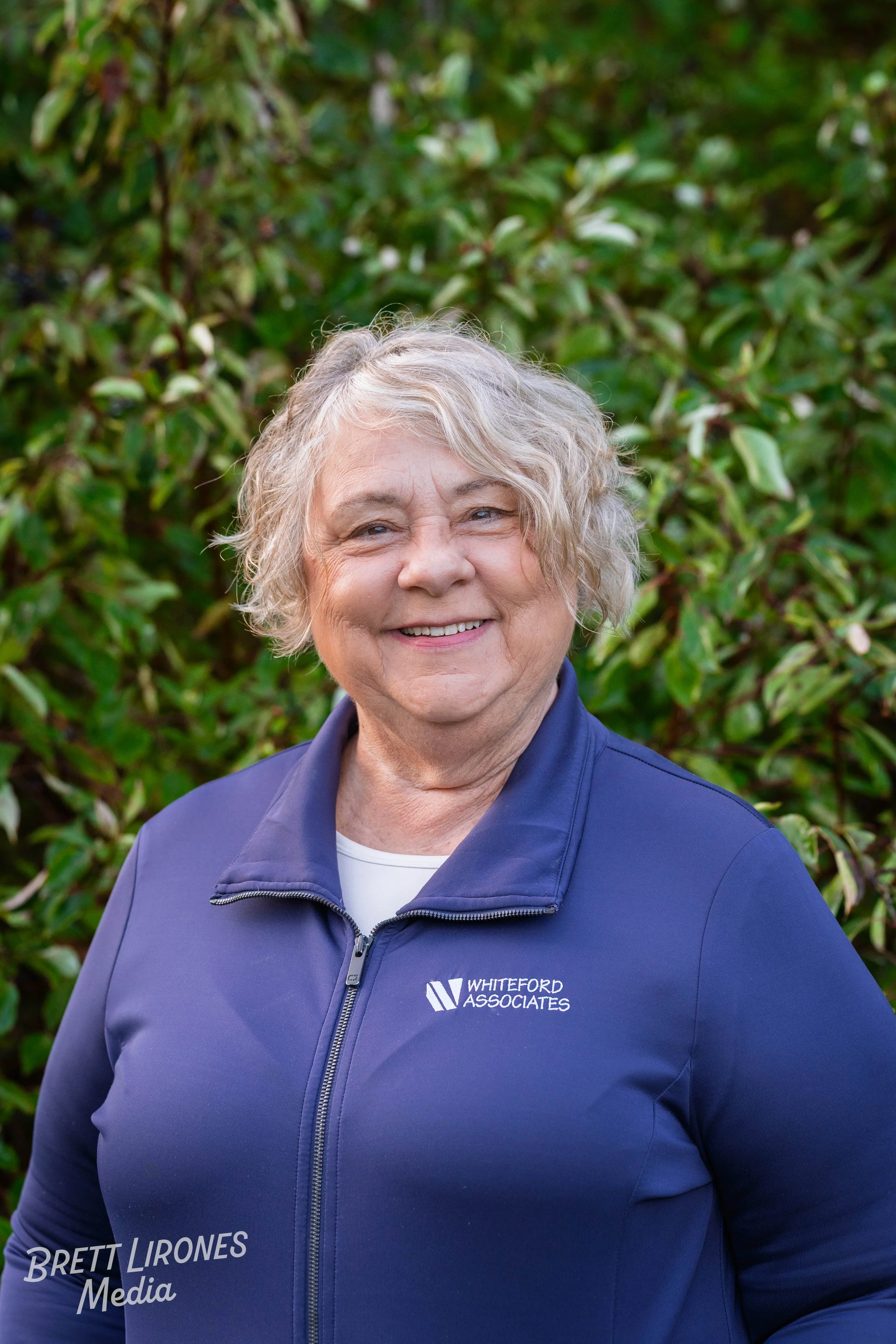 A smiling elderly woman with short, curly gray hair standing outdoors in front of green leafy bushes, wearing a navy blue jacket with 'Whiteford Associates' logo.