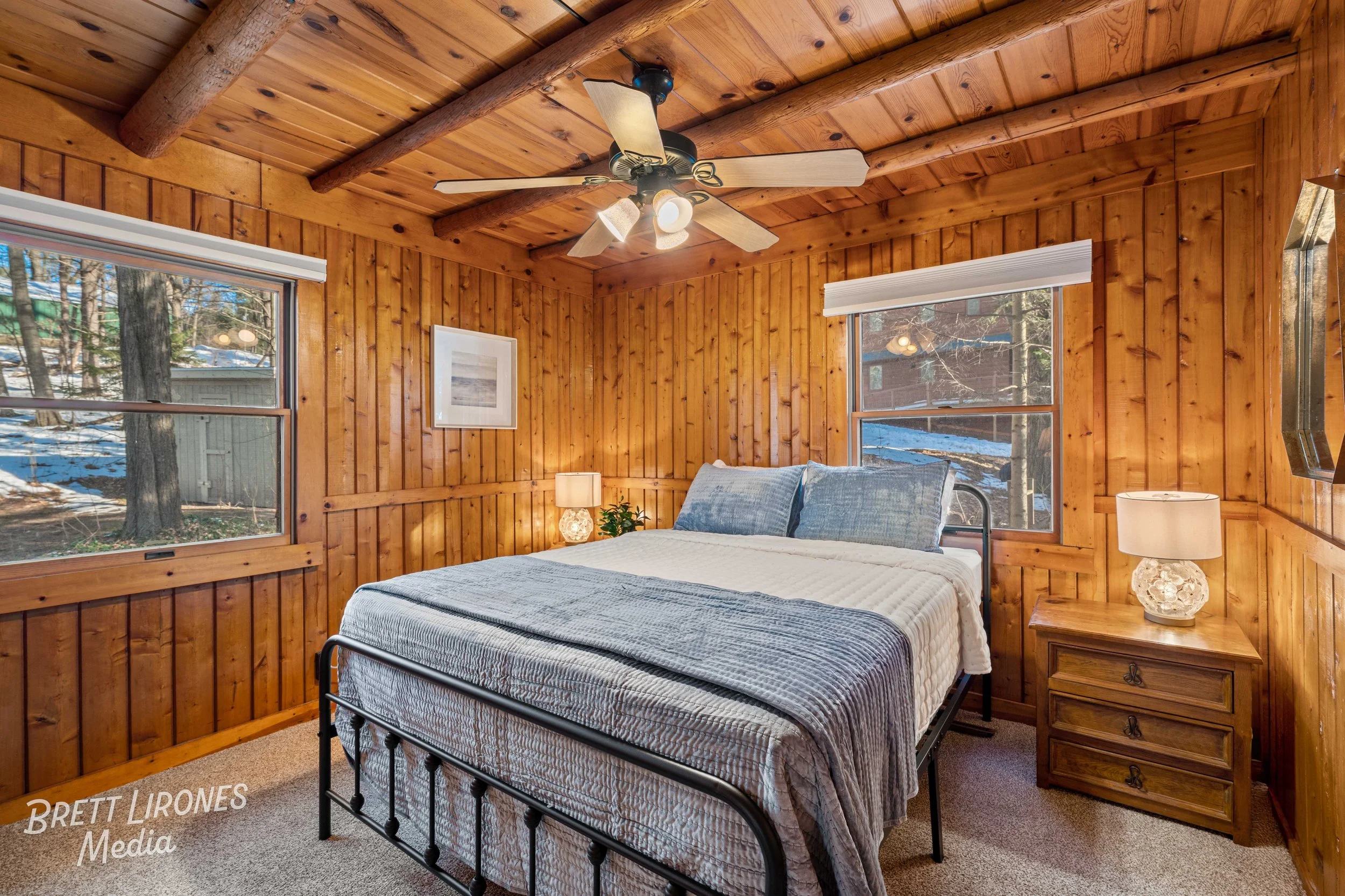 A cozy bedroom with wooden paneled walls and ceiling, two windows showing trees outside, a metal bed with blue pillows and a white blanket, a wooden nightstand with a lamp, and a ceiling fan.