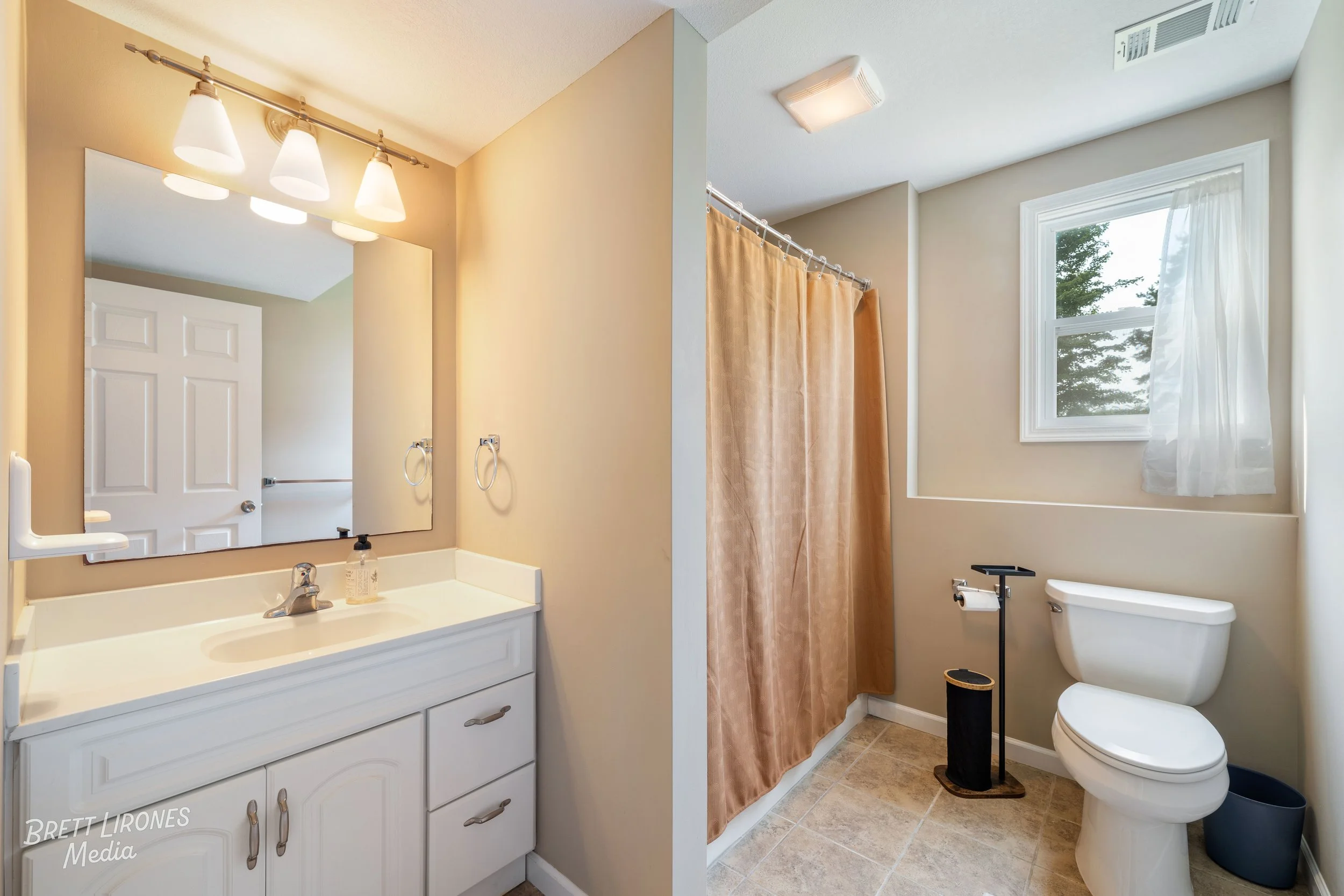 Bathroom with white vanity and mirror on the left, light beige walls, shower with beige curtain in the middle, and a white toilet near a window with white curtains on the right