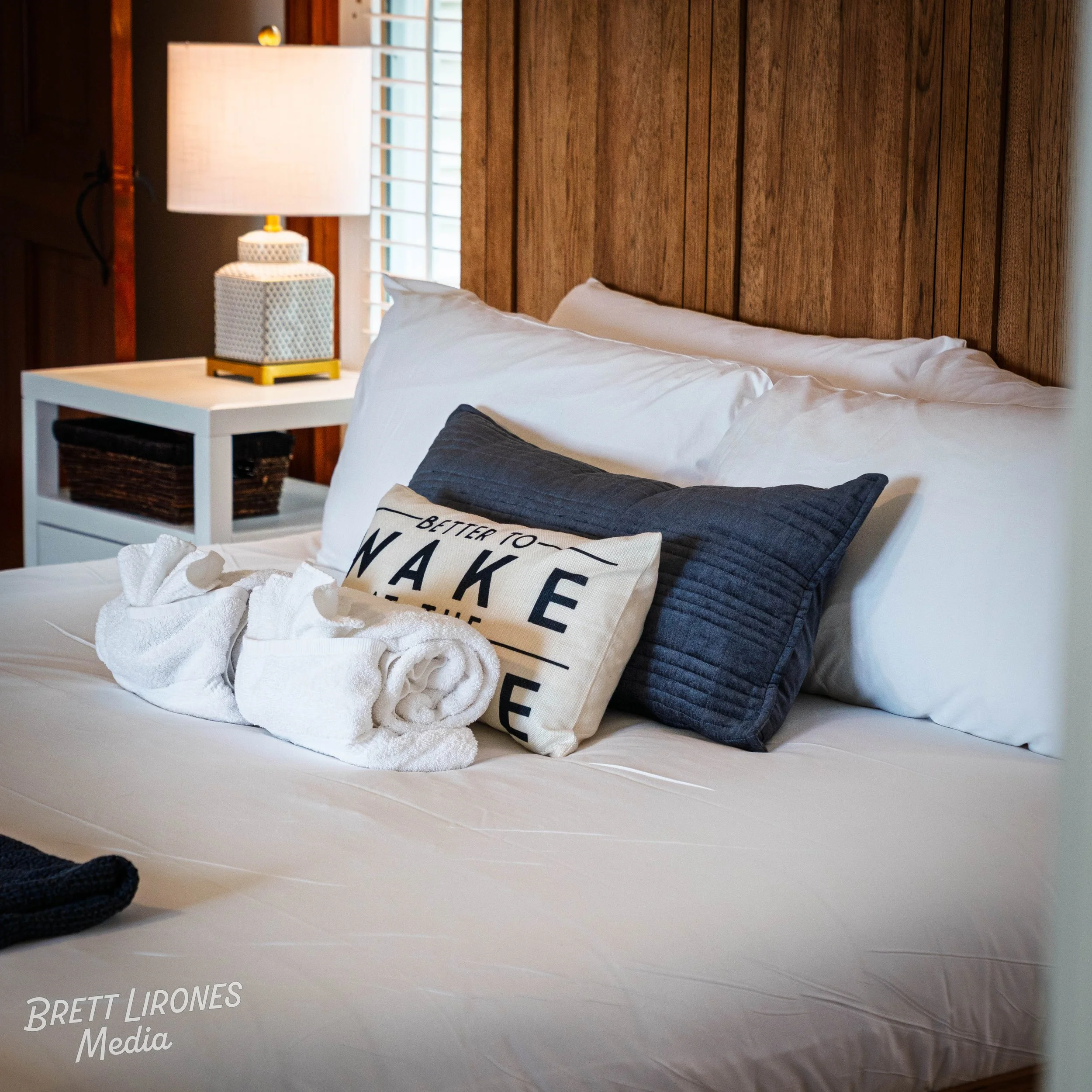A neatly made bed with white and dark gray pillows, a white towel, and a decorative pillow that says 'Better to make' in a cozy bedroom with wood paneling and a large window.