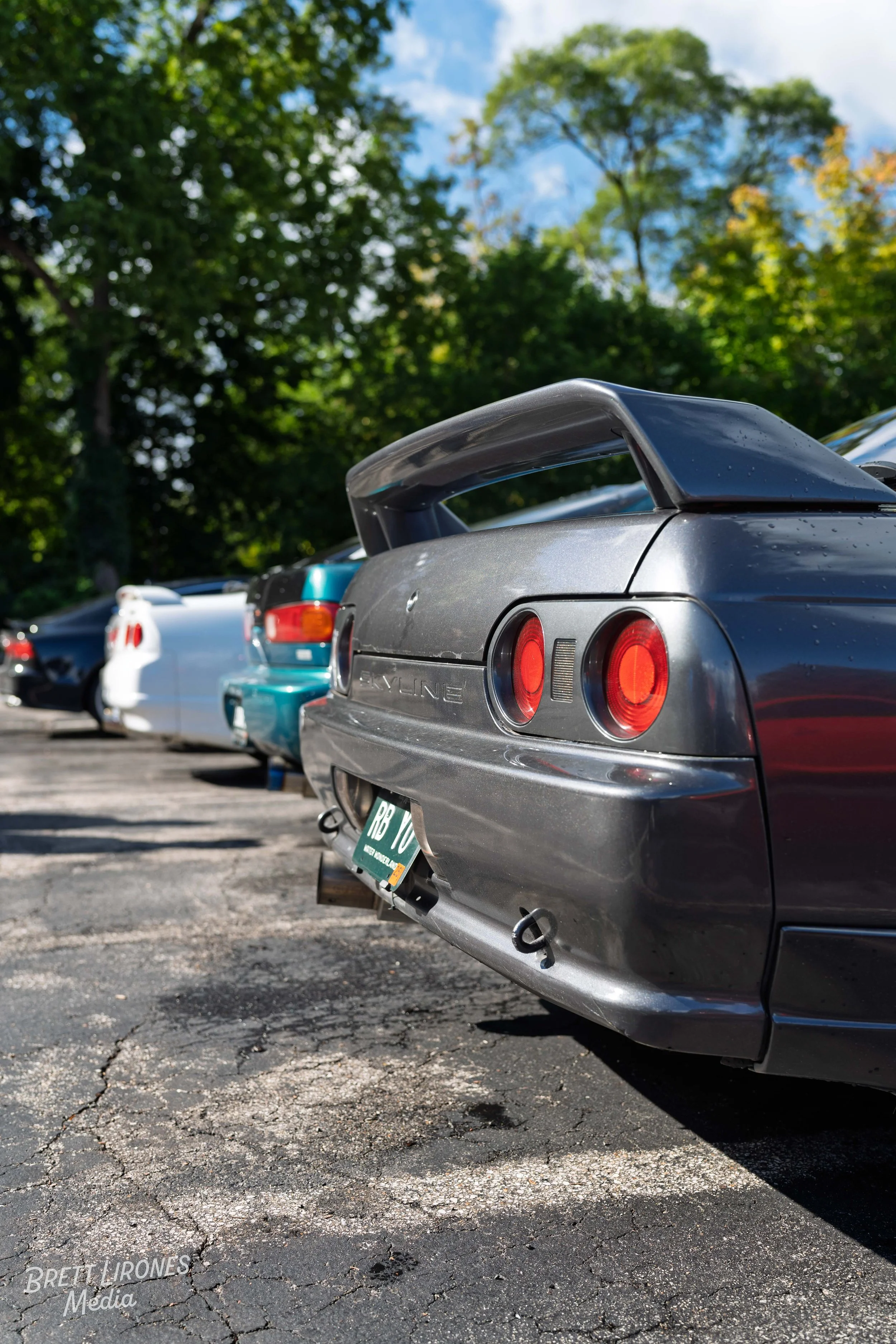Rear view of a gray Nissan 240SX sports car with a large spoiler, parked in a lot with other cars and surrounded by green trees.