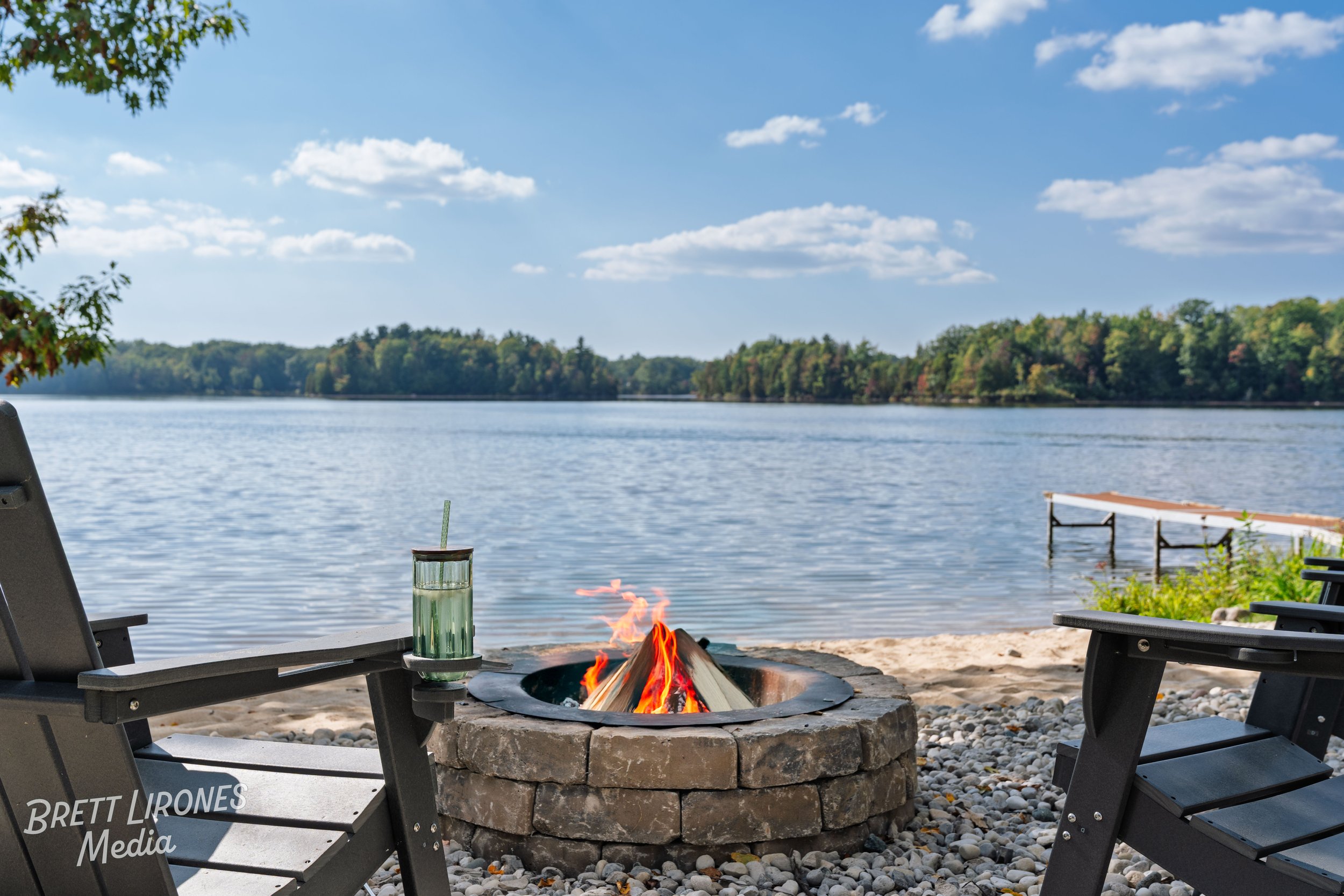 Beachside campsite with two lounge chairs, a fire pit with a burning fire, a water bottle with a straw on one chair, and a dock extending into a calm lake, with trees and a blue sky in the background.