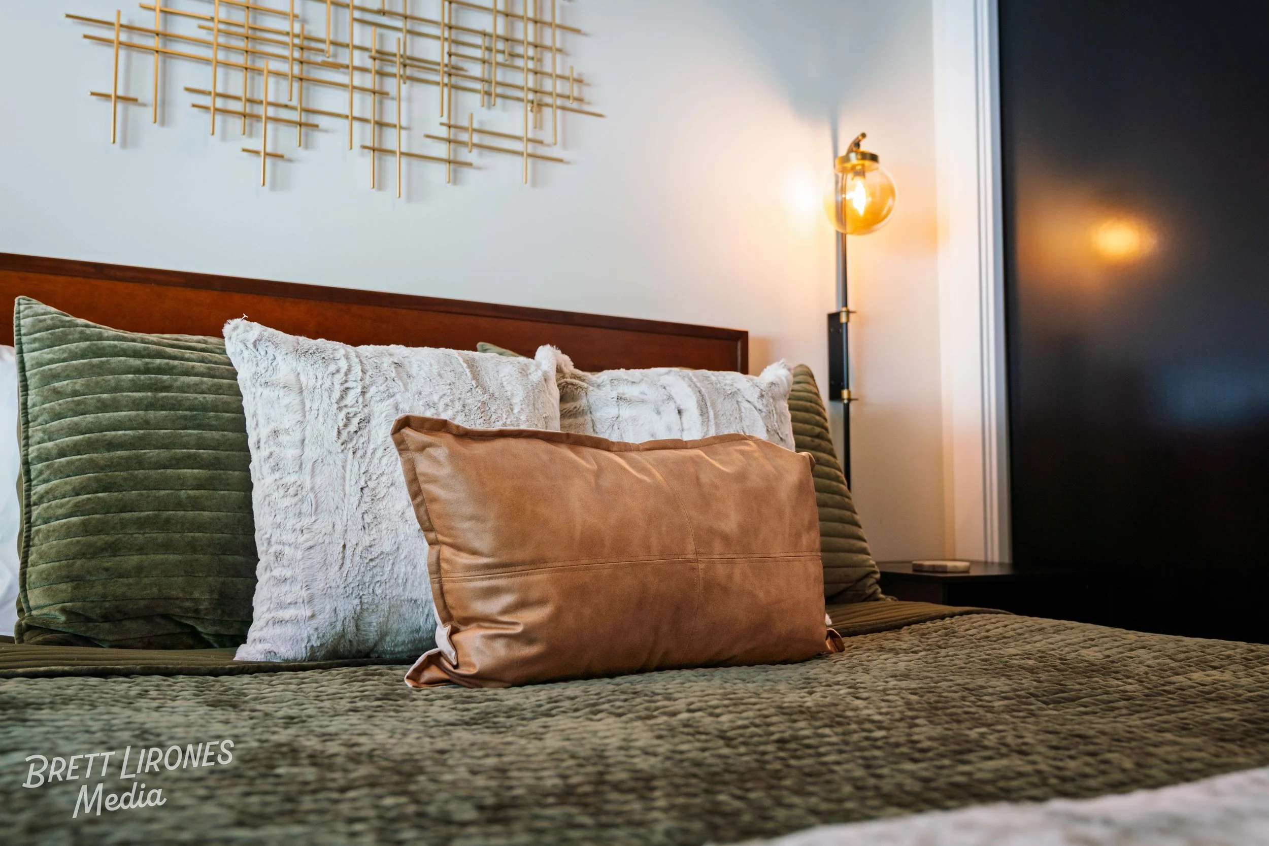 View of a neatly made bed with multiple pillows, including green, white, and tan pillows, in a modern bedroom with a wall-mounted light fixture and an abstract metal wall art piece.