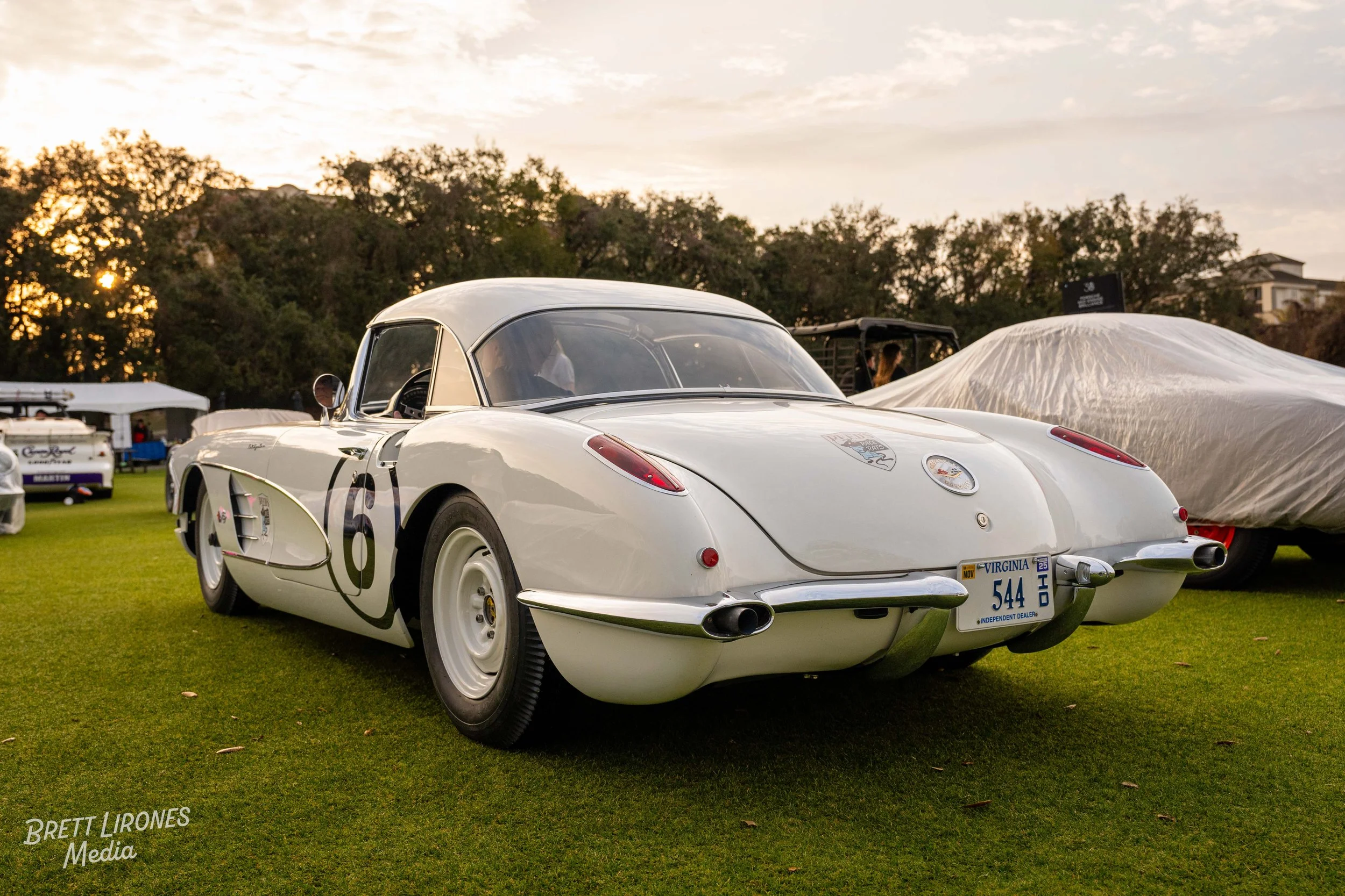 A classic white vintage race car with black racing number 6 on the side, parked on a grassy field during a car show at sunset.