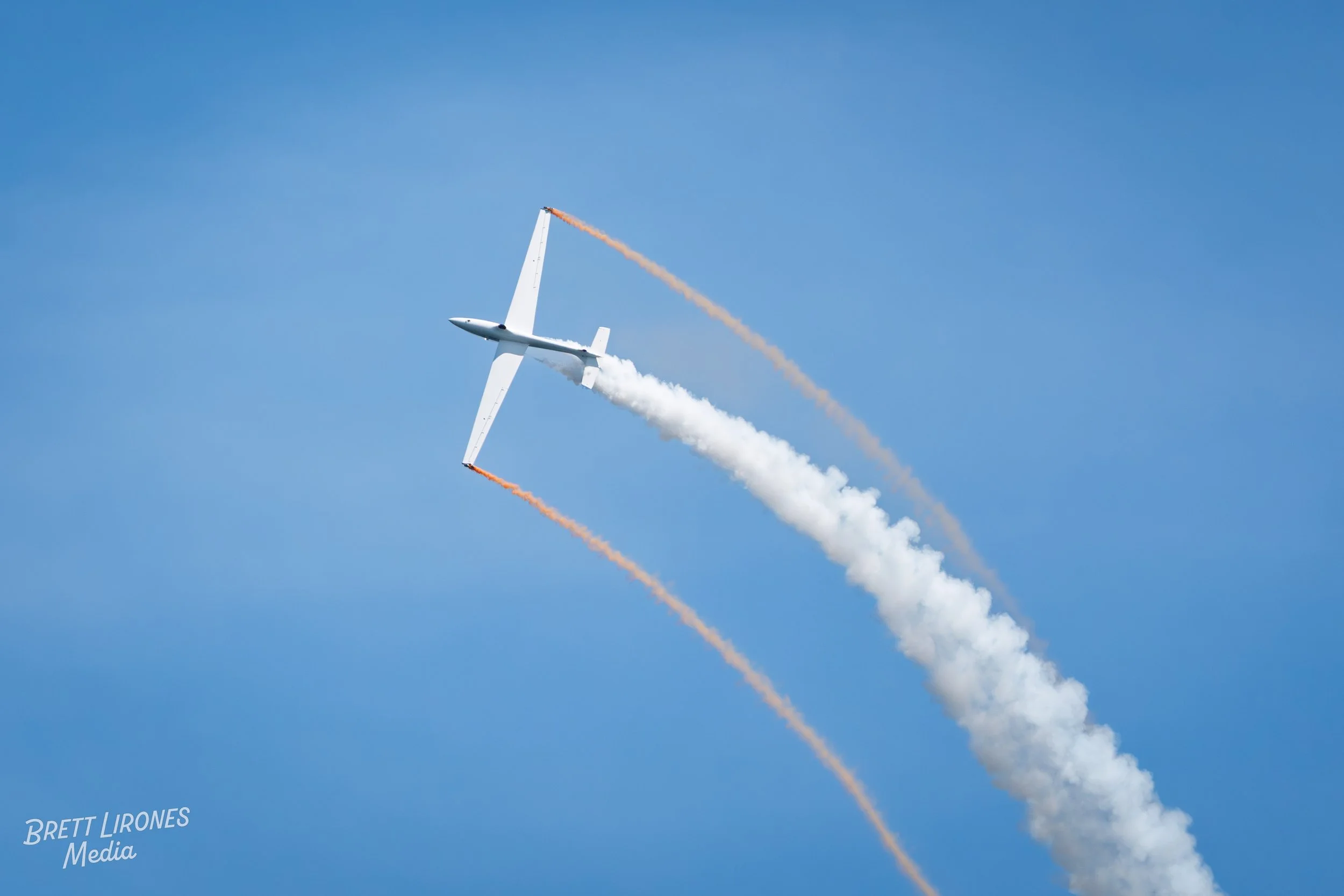 A white aerobatic aircraft performing a stunt in the sky, leaving white and orange smoke trails behind against a blue sky.