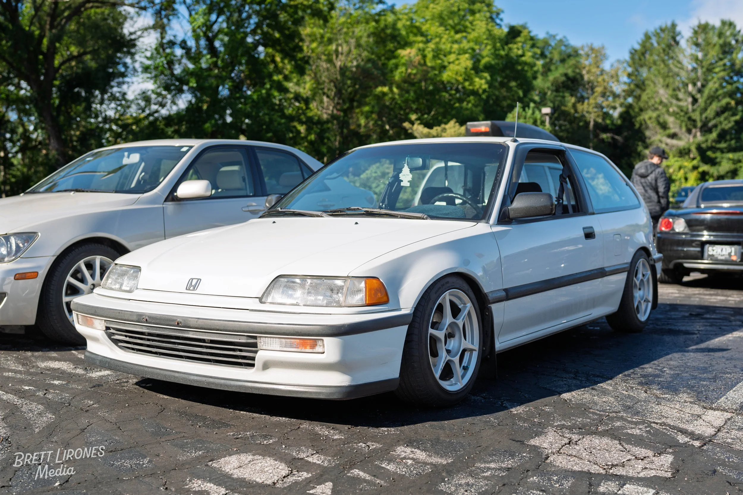 A white vintage Honda CR-X parked among other cars in an outdoor parking lot on a sunny day, with trees and a few people in the background.