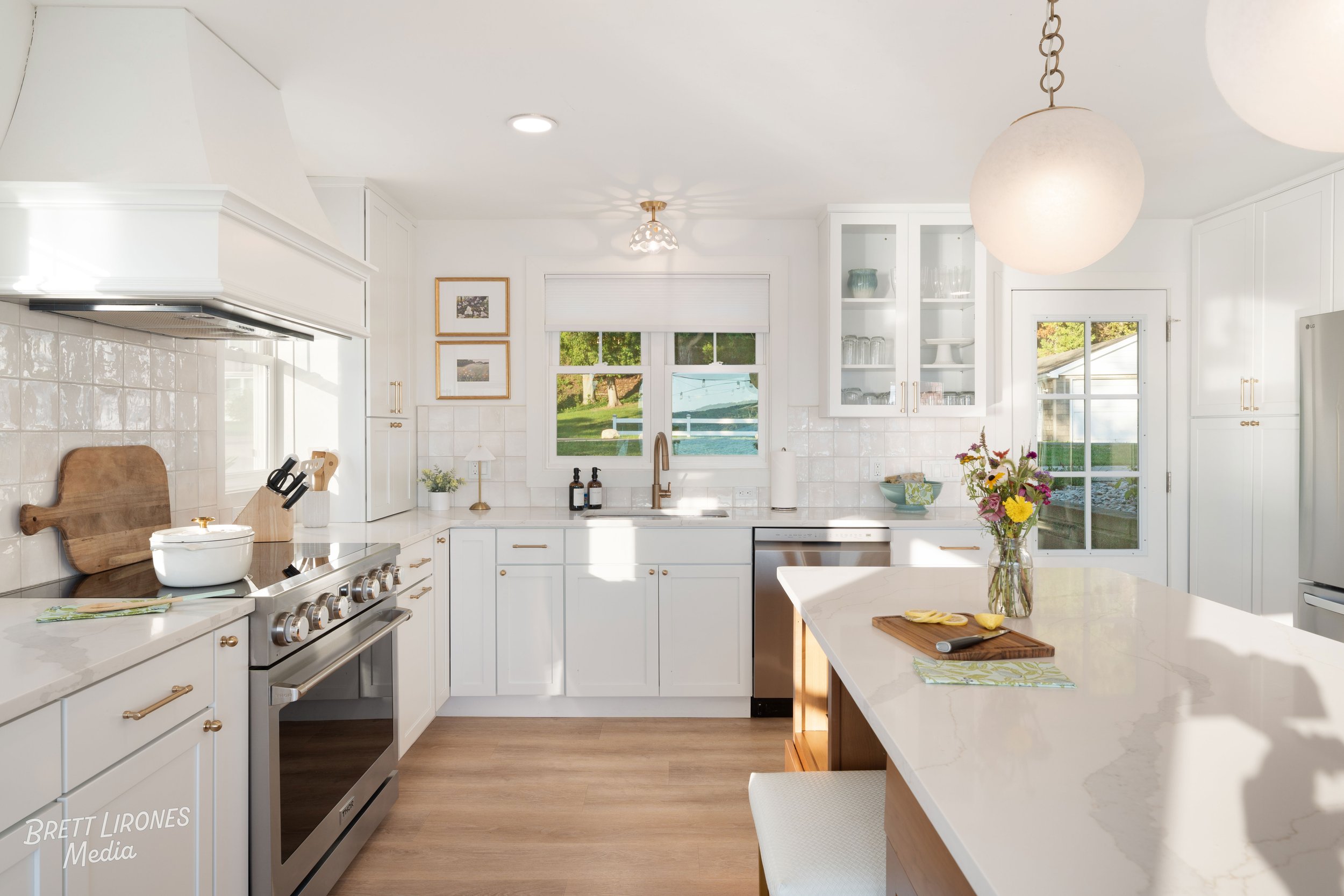 Bright, white kitchen with modern appliances, a large island with a vase of flowers, and windows showing greenery outside.