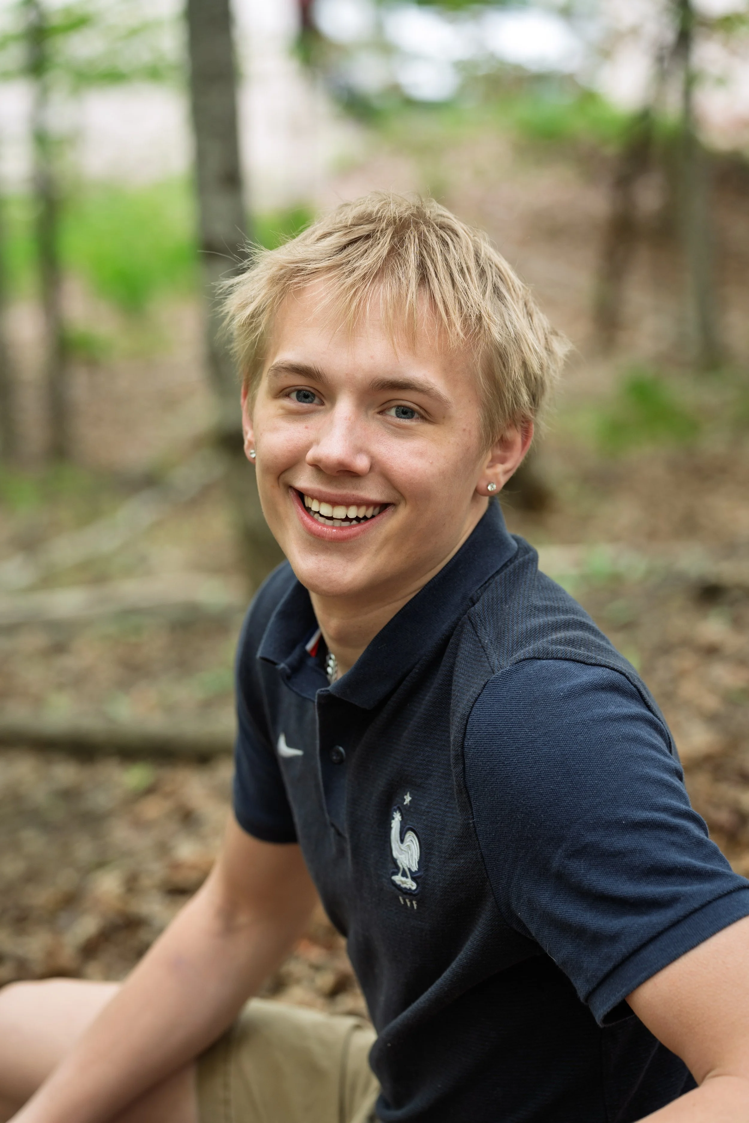 A young man with blond hair and blue eyes smiling outdoors in a wooded area, wearing a navy blue polo shirt with a rooster emblem and khaki shorts.
