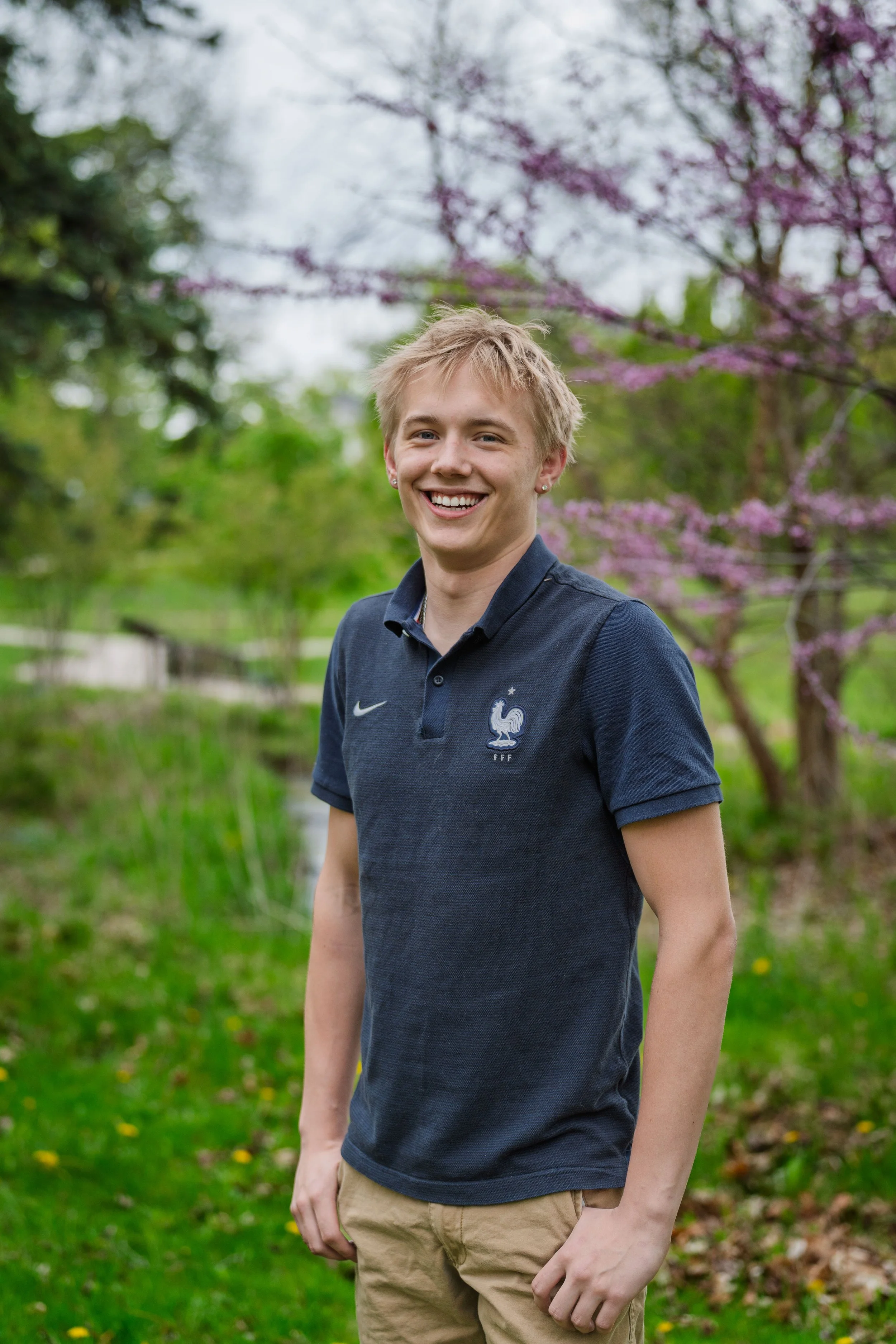 A young person with short blond hair smiling outdoors, wearing a navy Nike polo shirt with a France football emblem, beige pants, standing in a lush green park background with pink flowering trees.