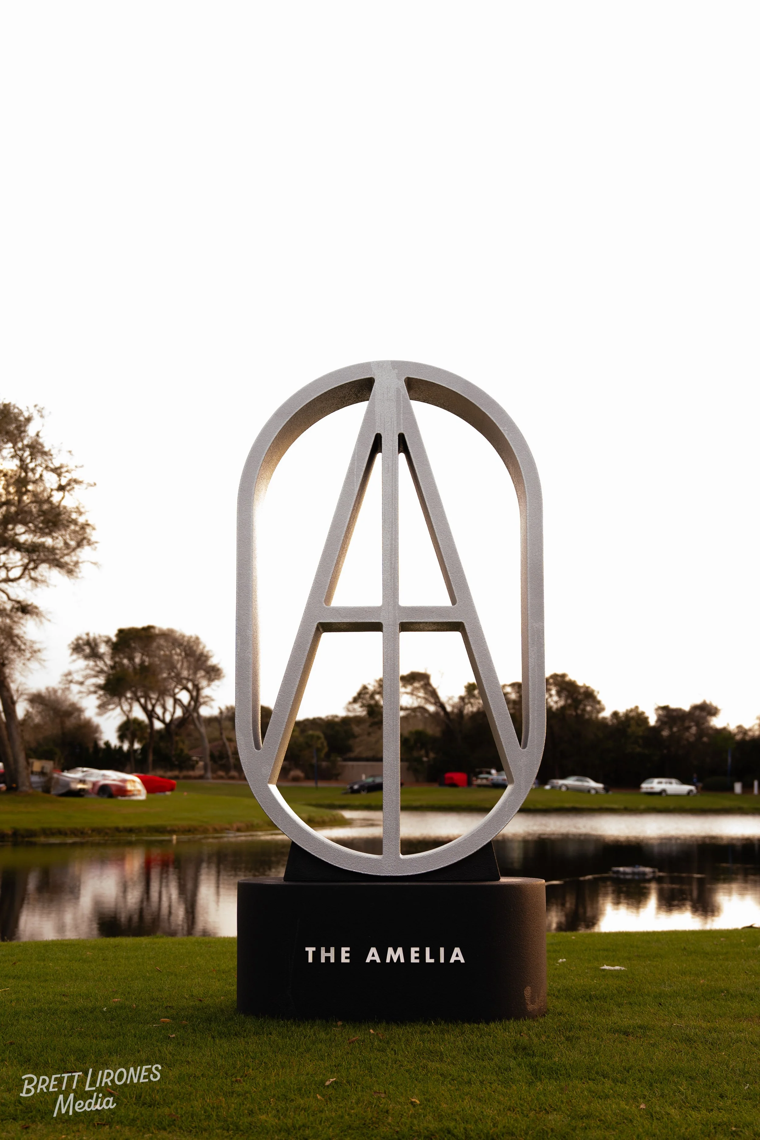 A sculpture of the peace symbol with the letter A inside, labeled 'The Amelia' on a black pedestal, set outdoors near a pond with trees and parked cars in the background during sunset.