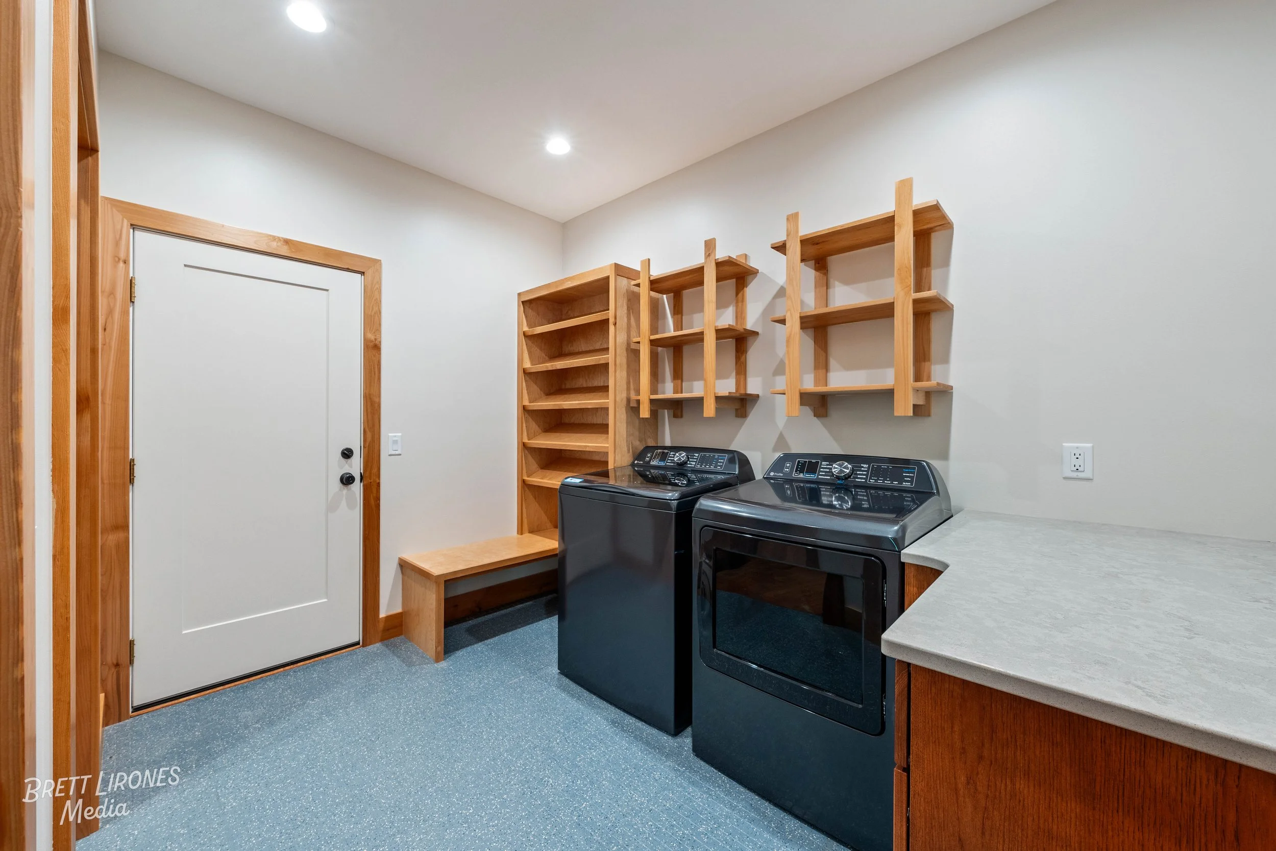 A laundry room with a white door, wooden shelves, a bench, a washing machine, a dryer, and a countertop, with a light-colored wall and ceiling.