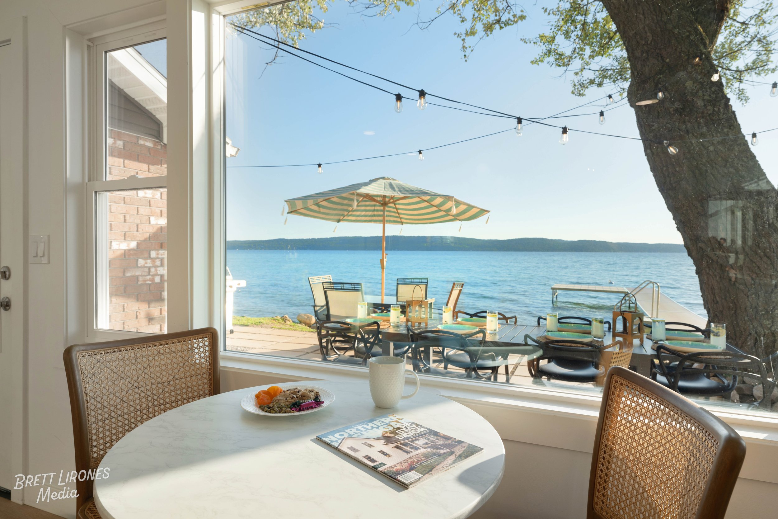 View of a lake seen from a dining area inside a house. The dining table has a magazine, a white mug, and a plate of food. Outside, a patio with a striped umbrella, chairs, and a dock extending into the water are visible, with a large tree on the righ