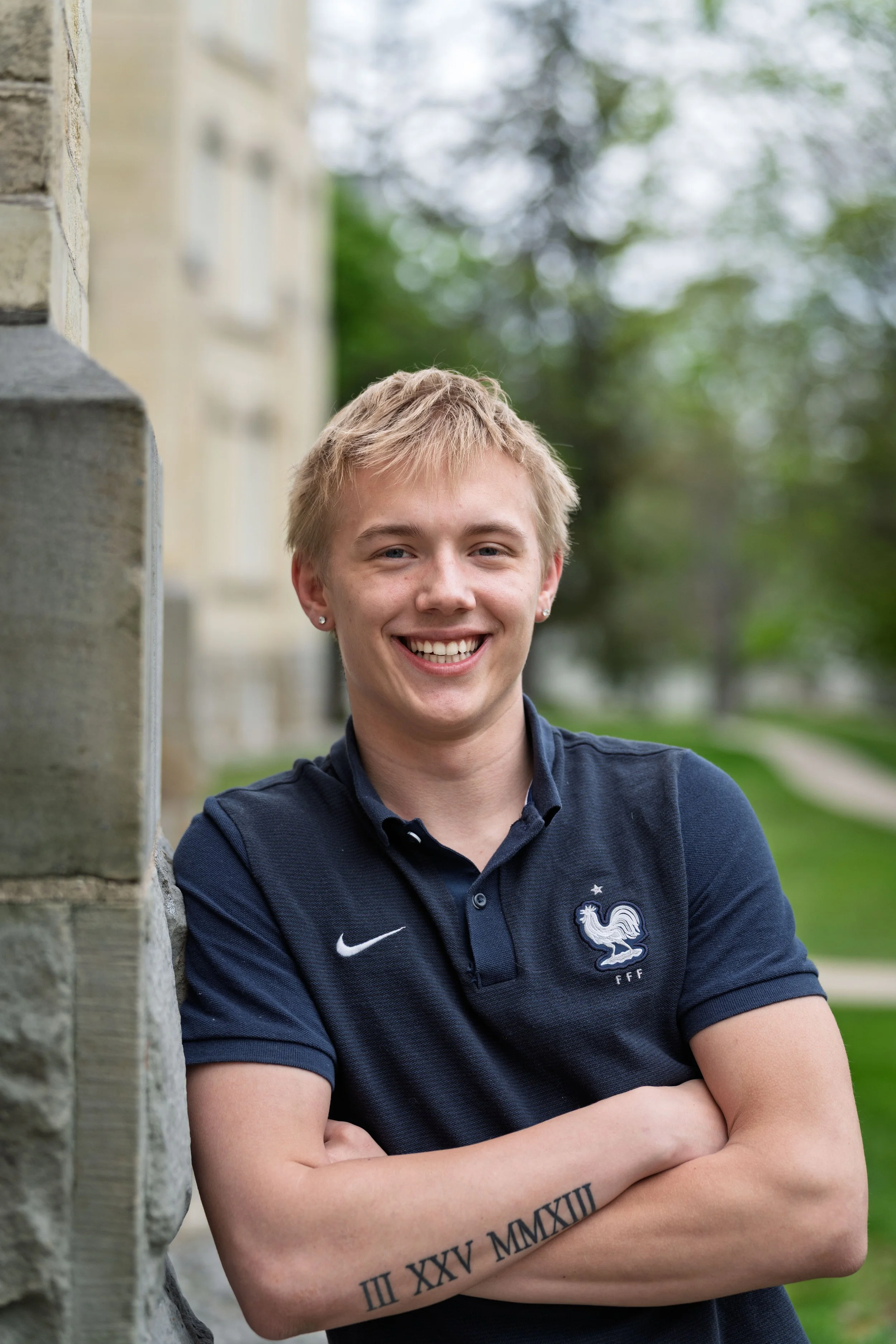 A young man with blonde hair, smiling, wearing a navy blue polo shirt with a French football emblem, standing outdoors with trees and a building in the background.