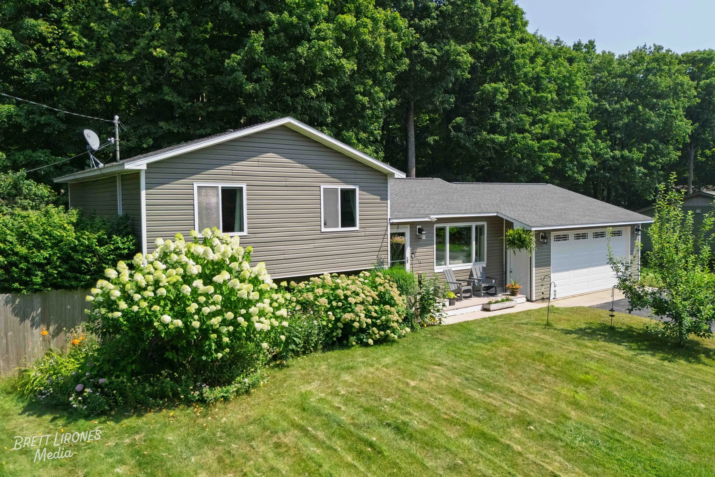 A single-story house with gray siding, white garage door, and front porch, surrounded by a green lawn, bushes, and trees, with a dense forest in the background.