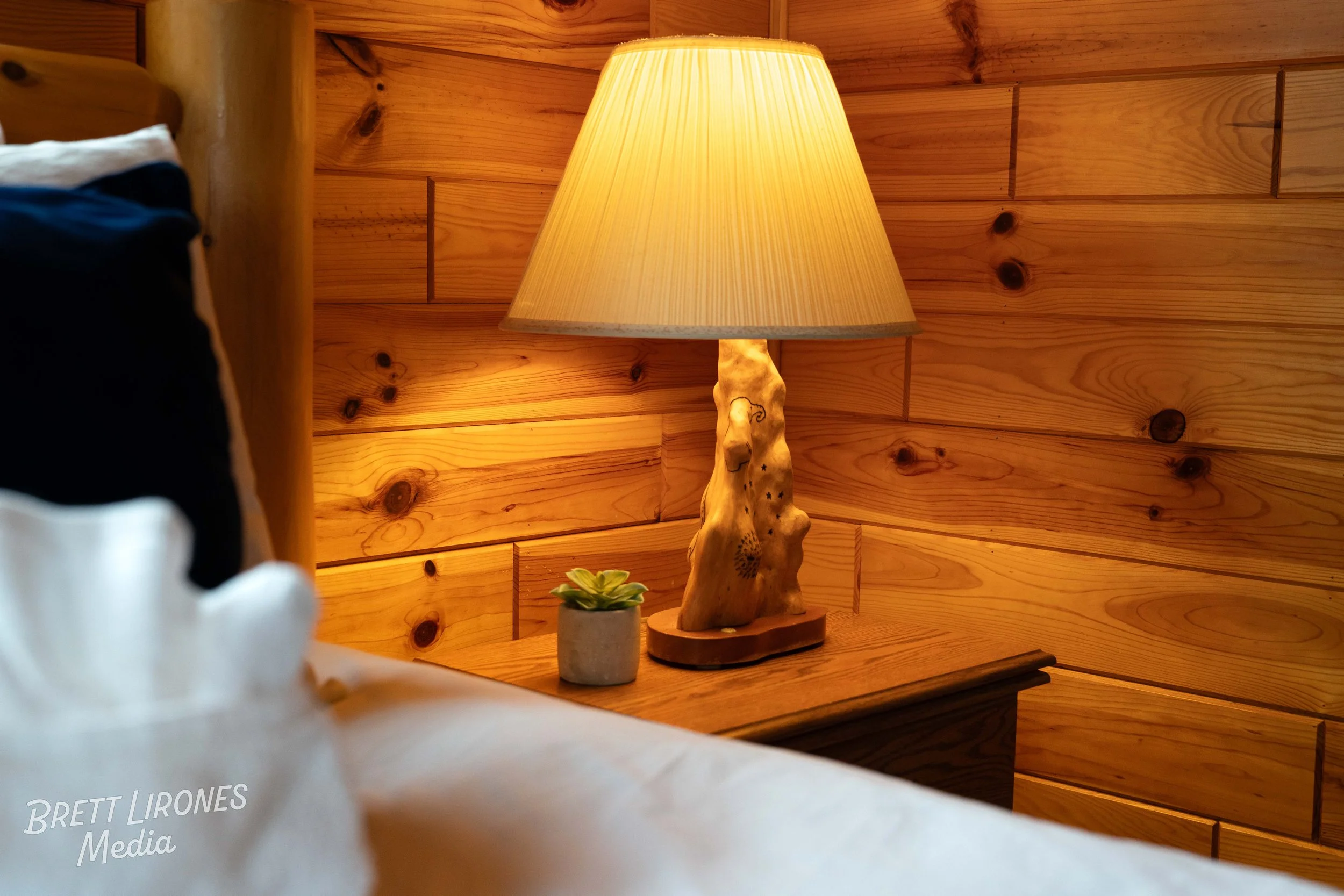 A wooden table beside a bed with a small potted plant and a lamp with a textured wooden base and a beige lampshade, set against a background of wooden wall paneling.