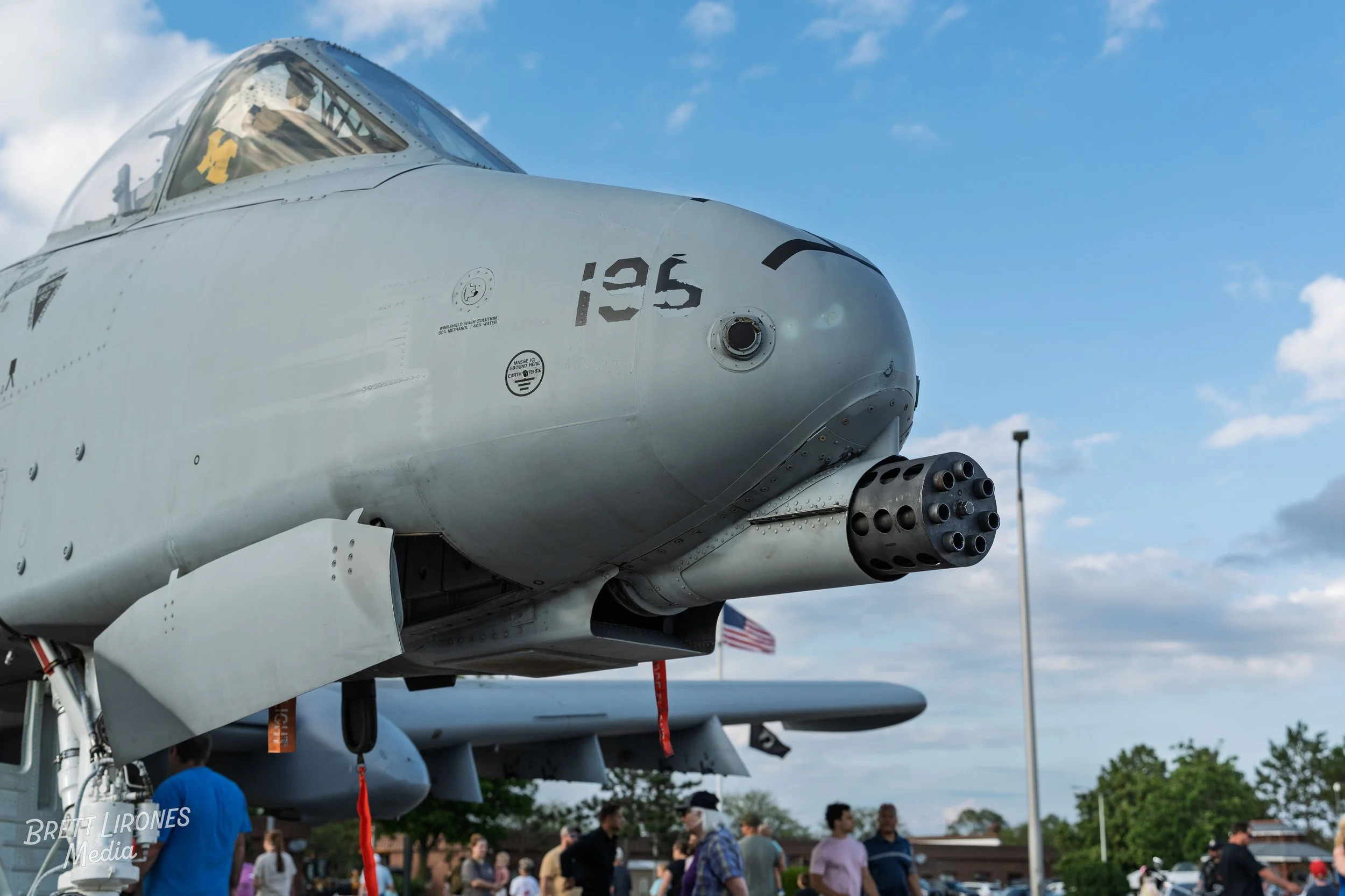 Front view of a military fighter jet on display outdoors with a crowd of people around it, under a partly cloudy sky.