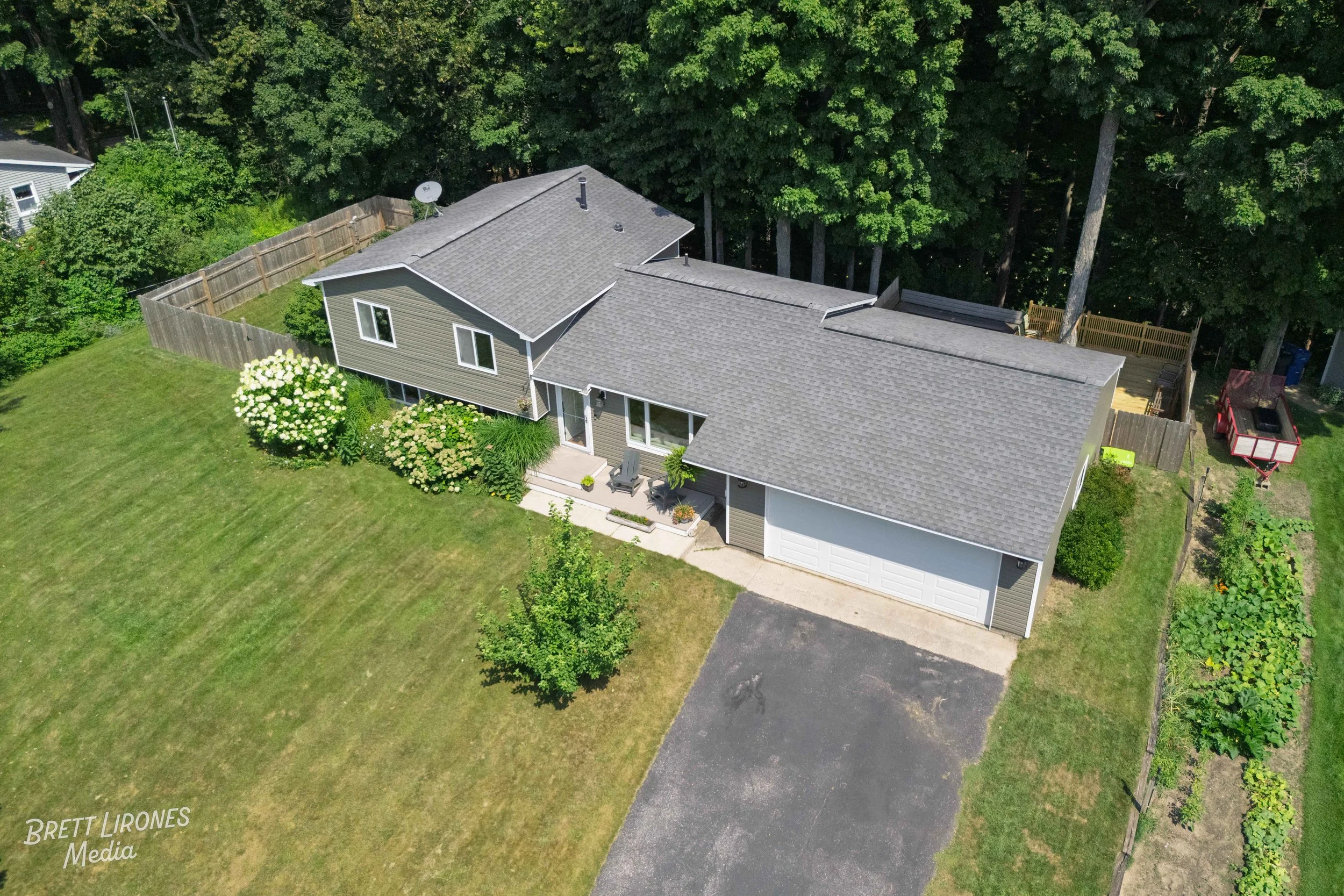 Aerial view of a modern suburban home with a large backyard, surrounded by trees, with a driveway, a small front porch with chairs, a garden area, and fenced yard.