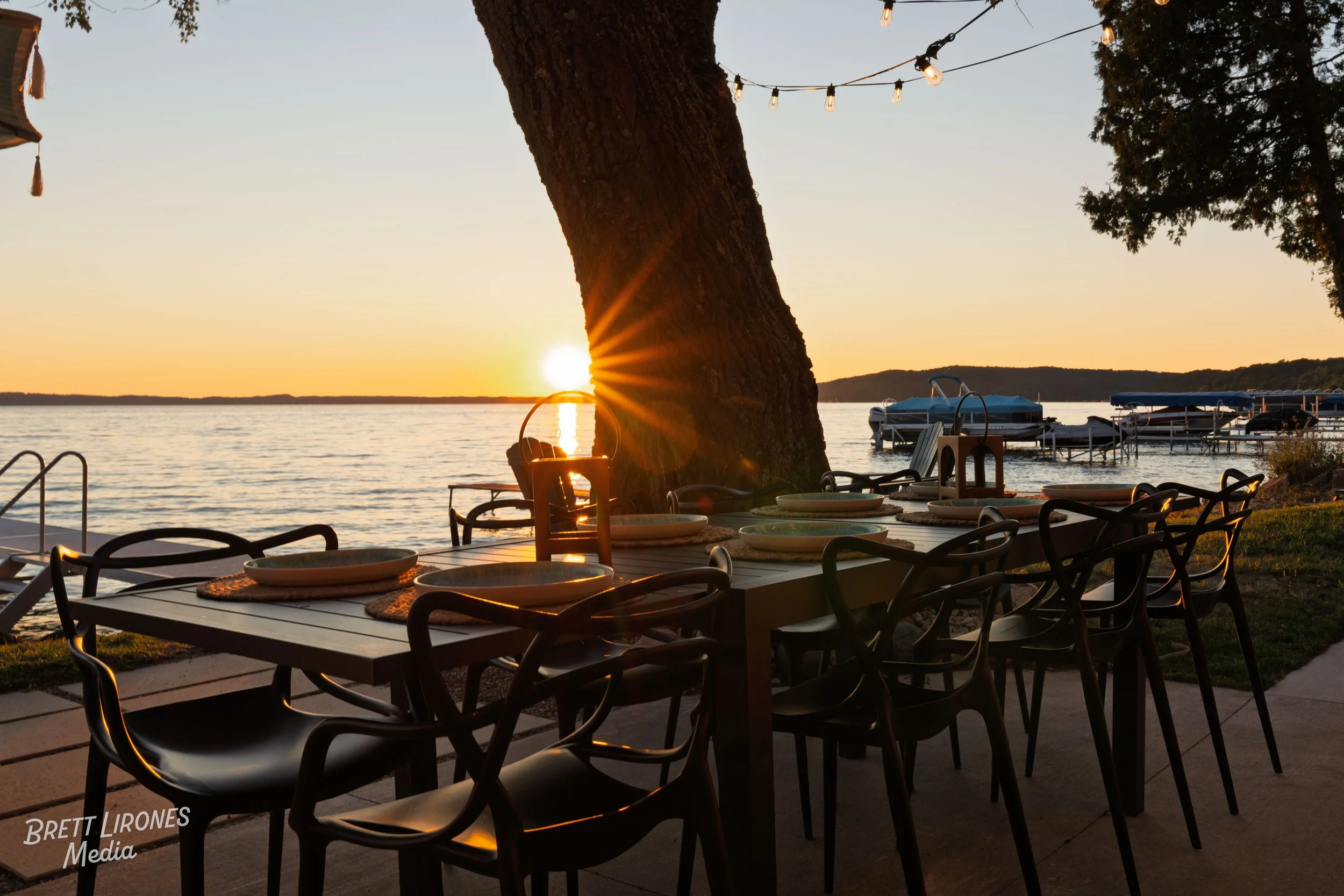 Outdoor dining table set near a lake at sunset, with string lights overhead, boats in the water, and a large tree in view.