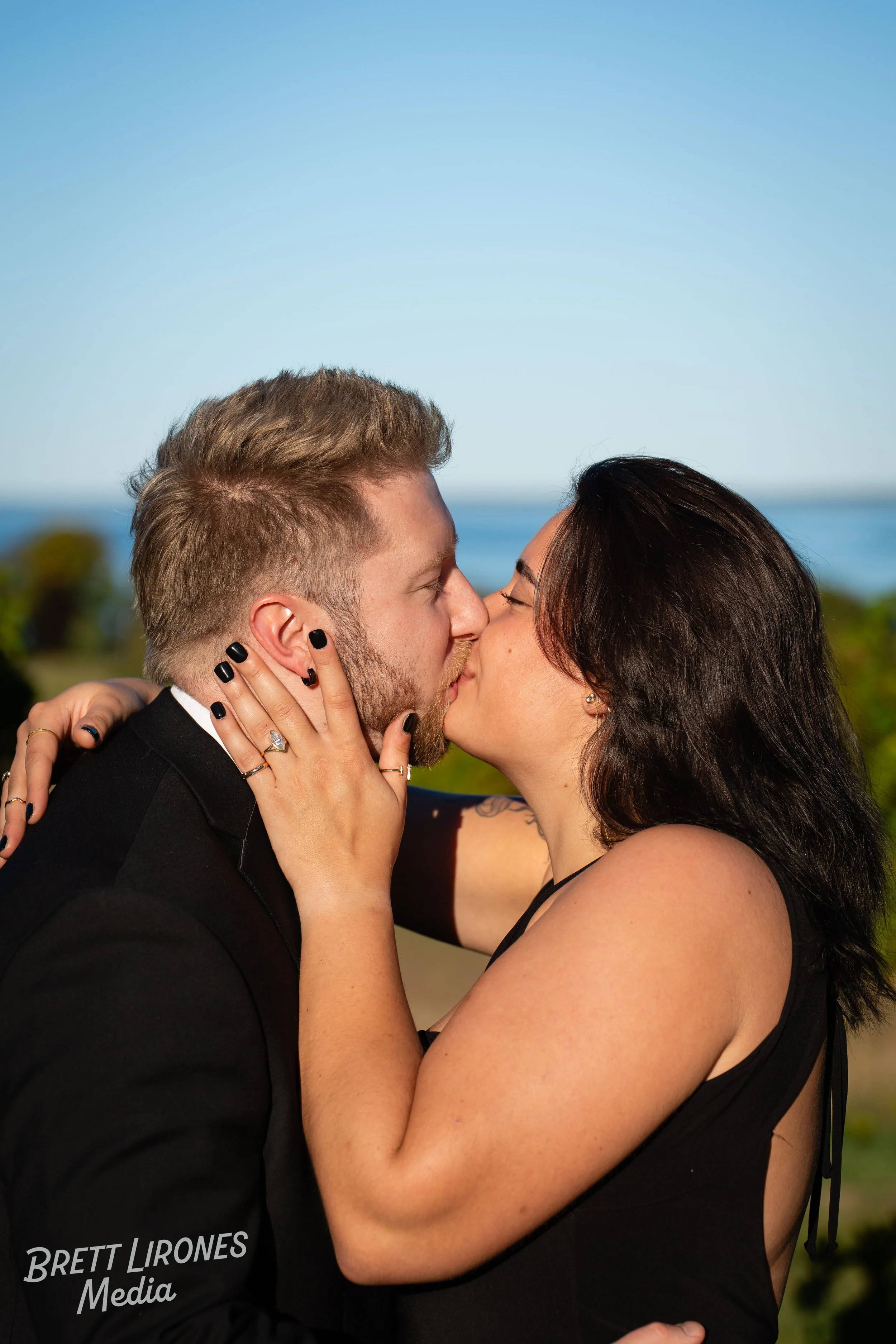 A couple is kissing outdoors with a scenic ocean background, the woman holding the man's face with her left hand.