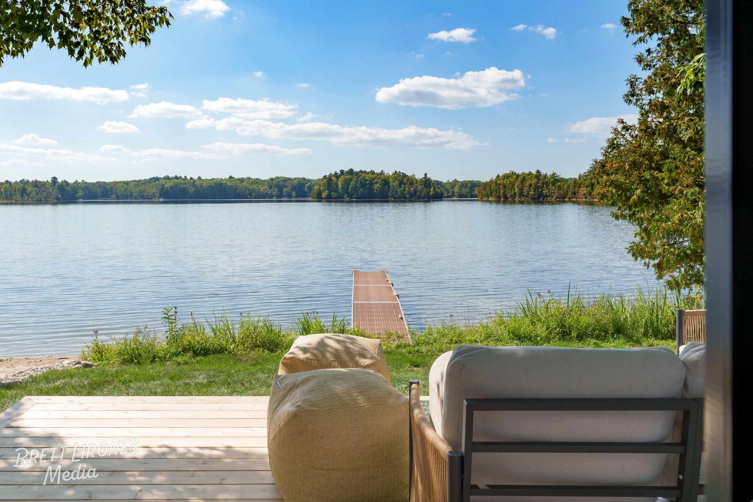 View of a calm lake with a dock extending into the water, seen from a porch with outdoor furniture, trees, and a blue sky with clouds.