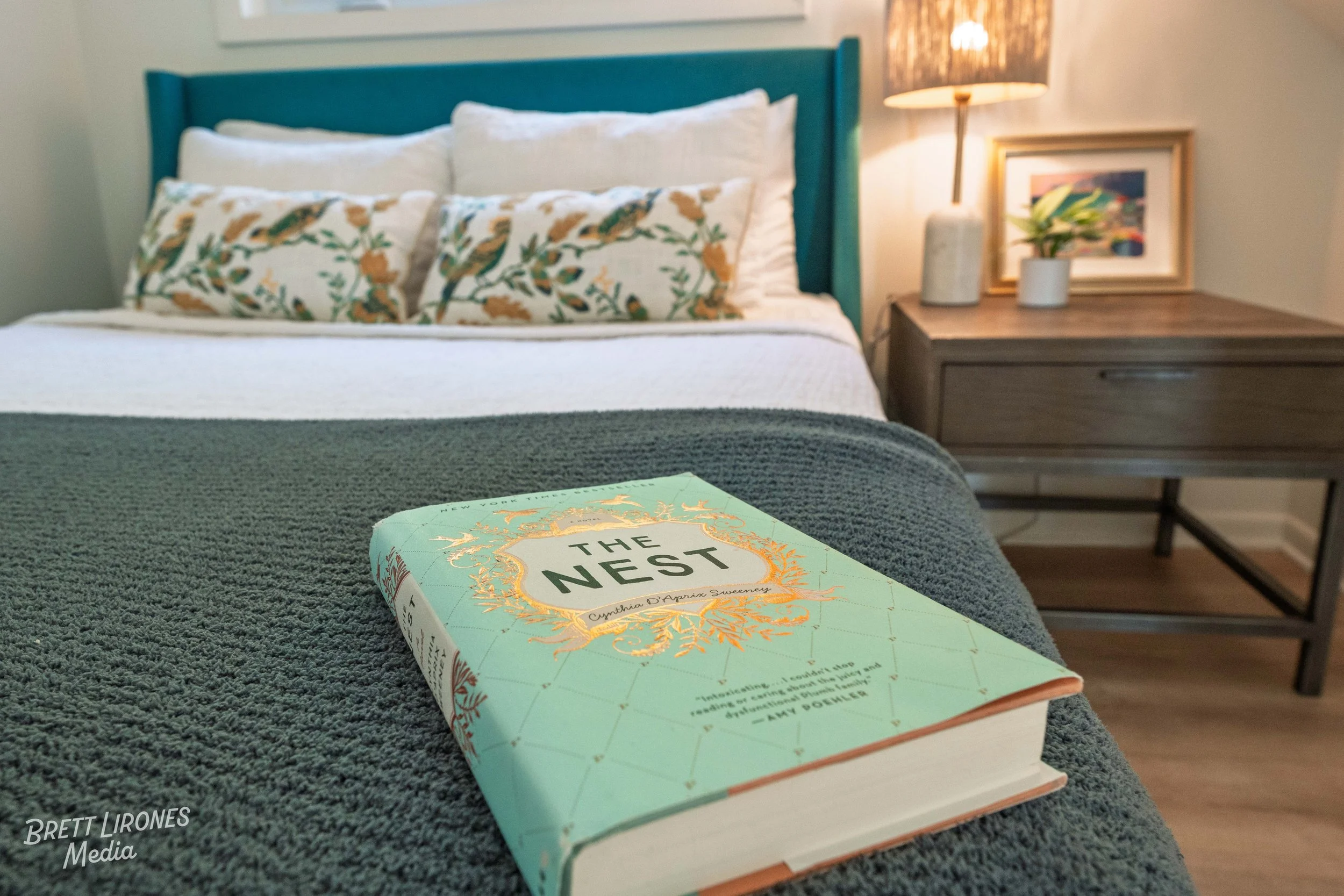 A hardcover book titled 'The Nest' resting on a gray comforter in a bedroom. The bed has two white pillows and a decorative pillow with a floral design. To the right, there is a nightstand with a lamp, a framed picture, and a potted plant.