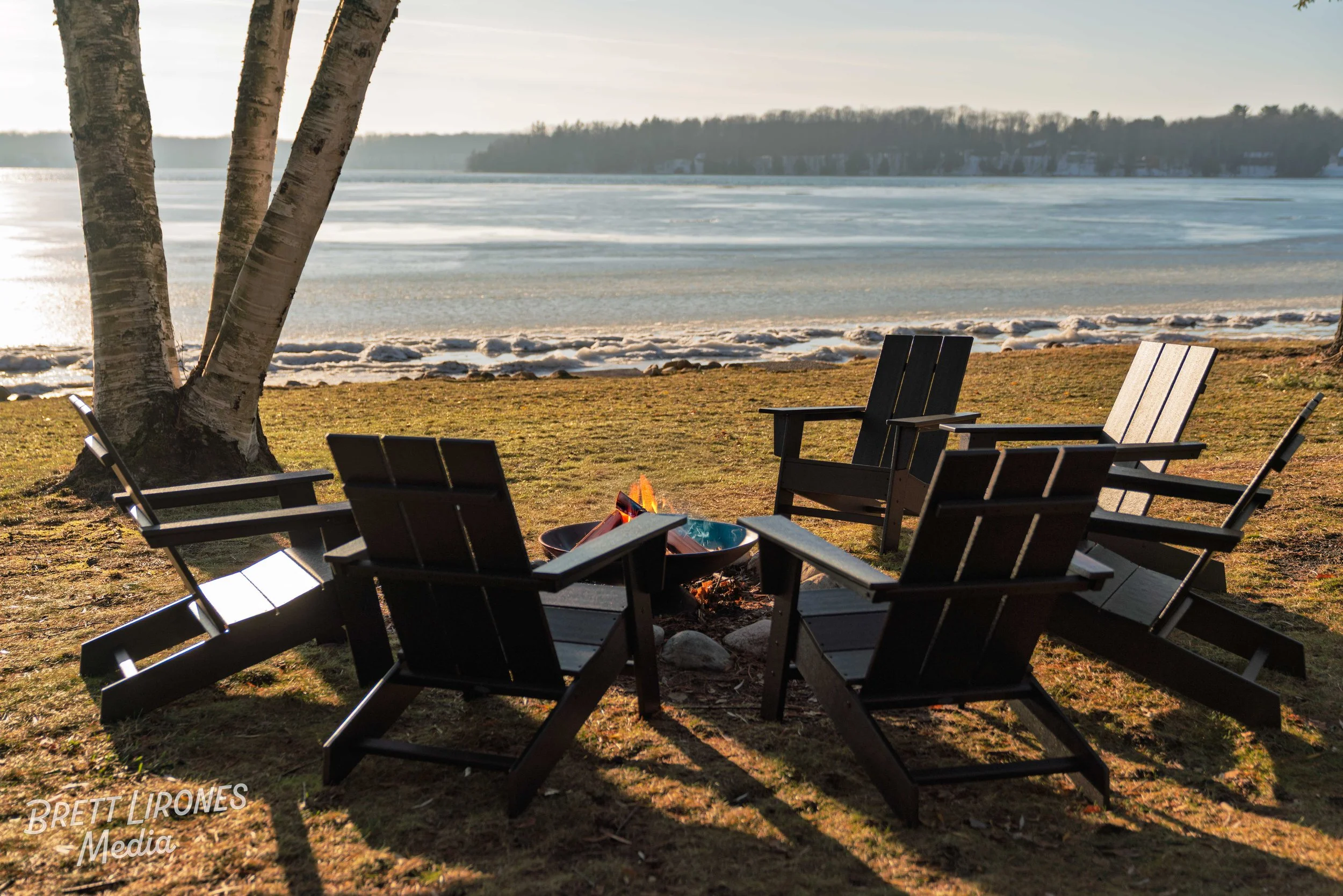 Six Adirondack chairs arranged in a semi-circle around a fire pit on a grassy lakefront, with a frozen lake and distant shoreline with trees in the background, under a mostly clear sky.