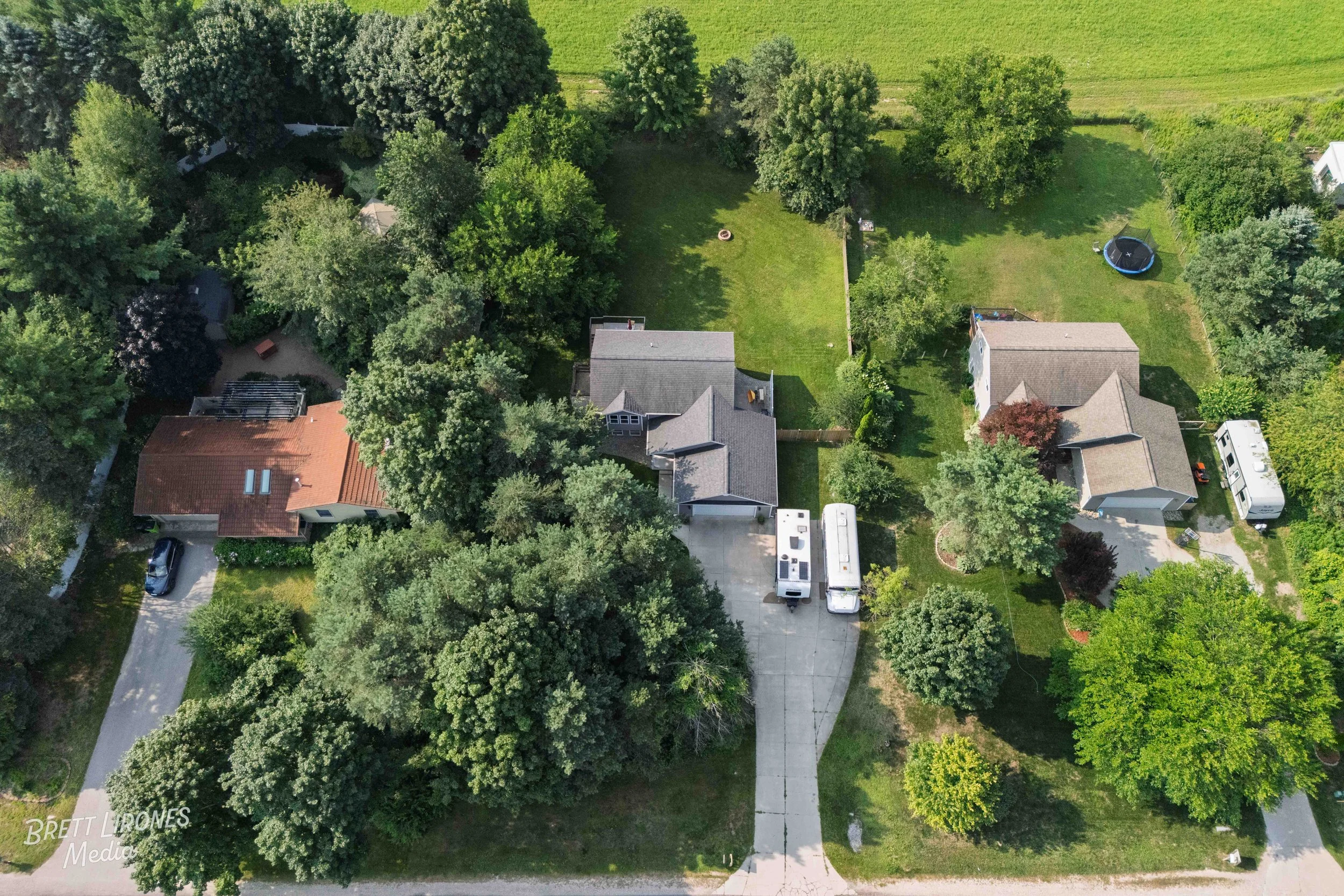Aerial view of a residential neighborhood showing three houses with backyards, trees, and a driveway with parked recreational vehicles.