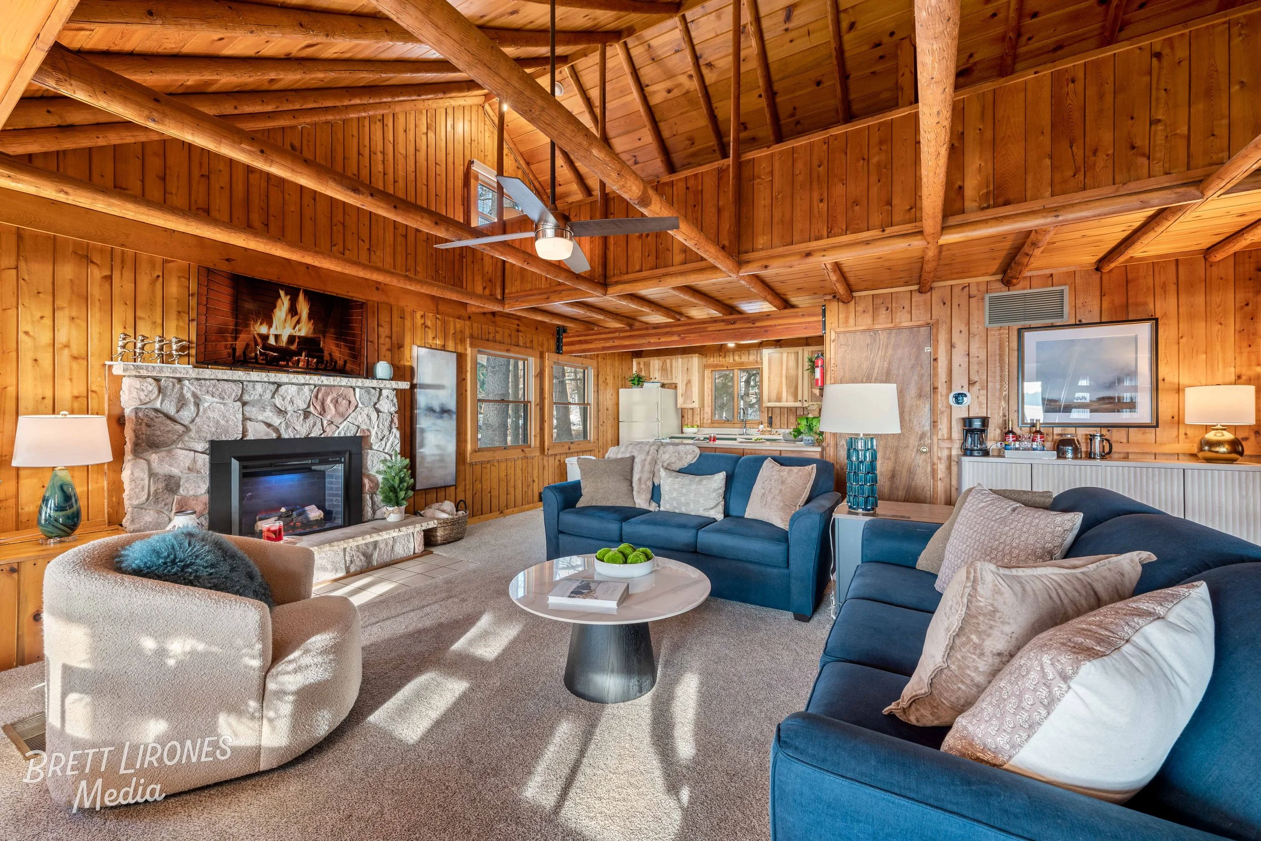 A cozy living room with wood-paneled walls and ceiling, featuring a stone fireplace with a fire burning, blue sofas with various pillows, a round coffee table with green apples, an armchair with a fuzzy pillow, and a kitchen area in the background.