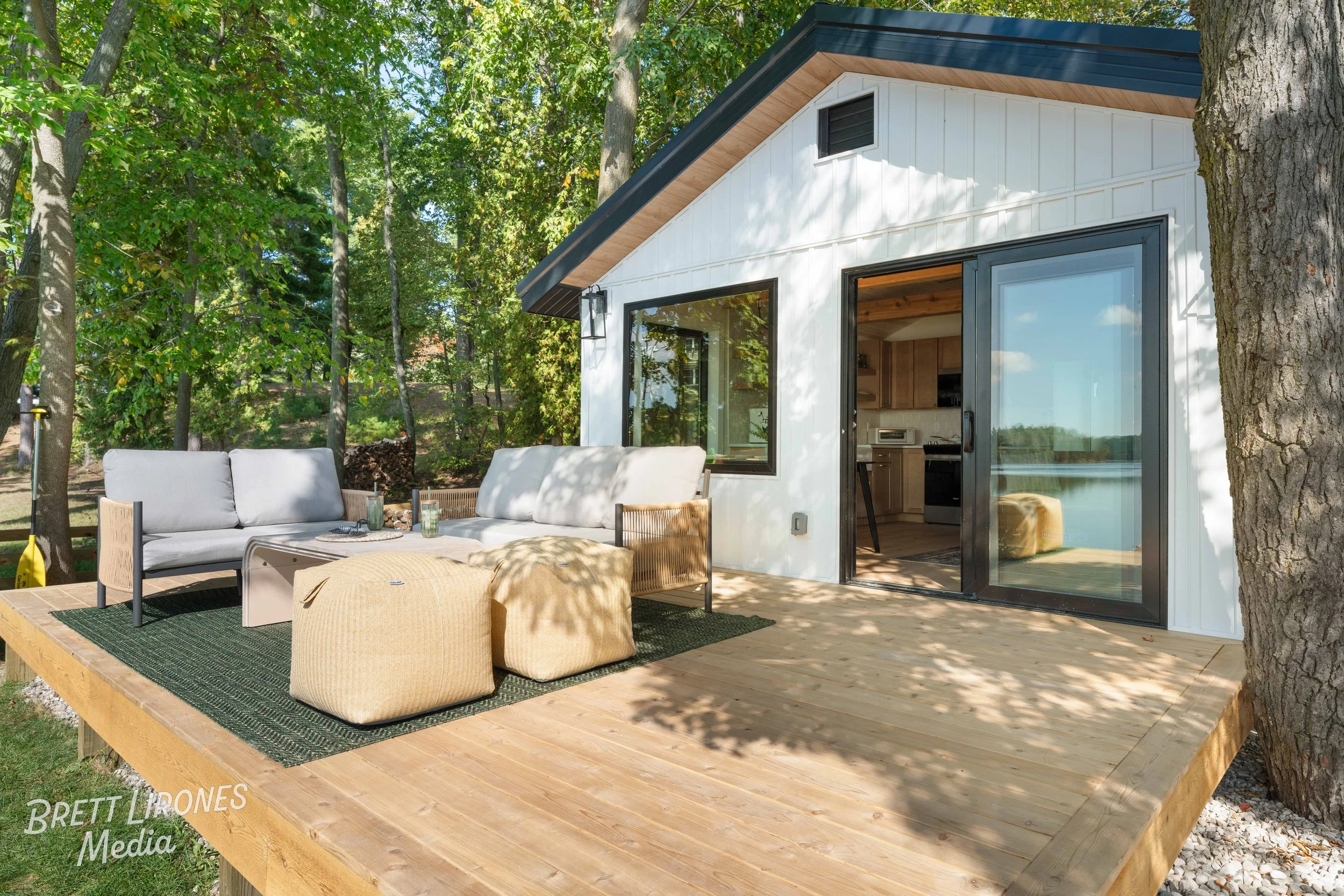 A wooden deck outside a white house with black trim, surrounded by trees, with outdoor furniture including a cushioned sofa, two beige poufs, and a coffee table, overlooking a body of water in the background.