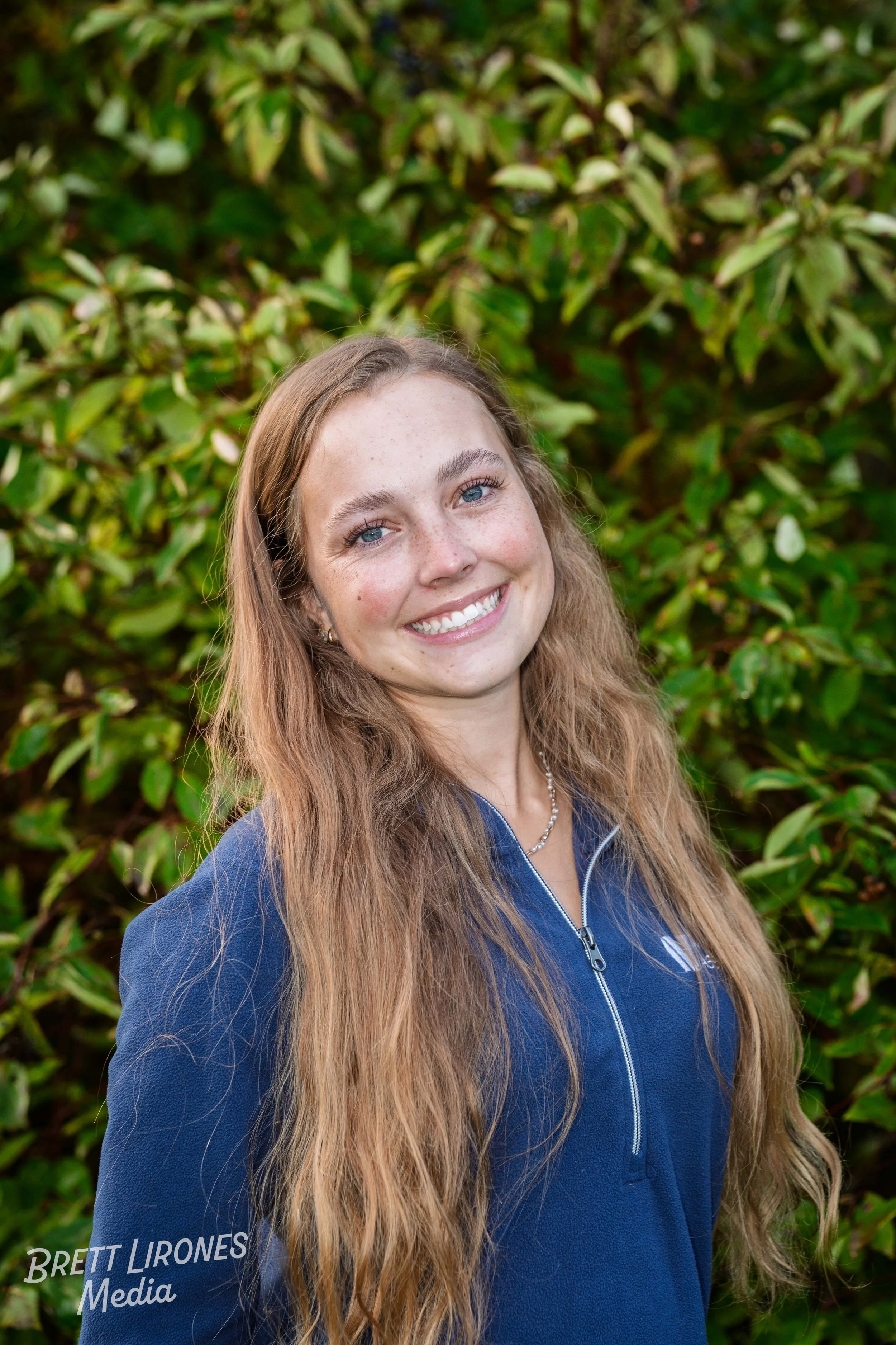 A young woman with long, wavy, light brown hair and blue eyes, smiling outdoors in front of green foliage, wearing a blue zip-up jacket and a silver necklace.