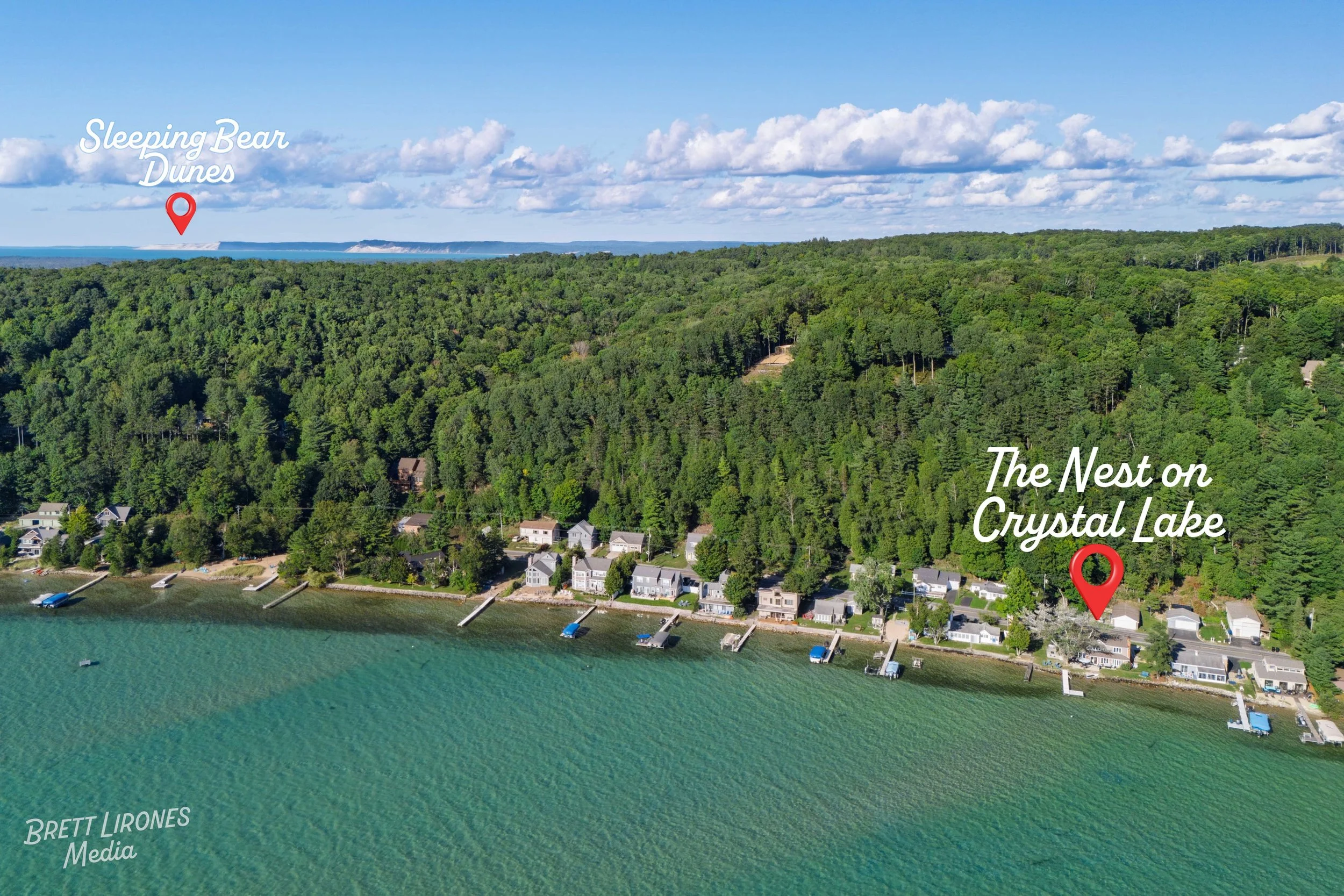 Aerial view of lakeside homes along Crystal Lake, with green forested hills in the background. Labels indicate 'Sleeping Bear Dunes' near the horizon and 'The Nest on Crystal Lake' next to a house. The water is turquoise and several docks extend into