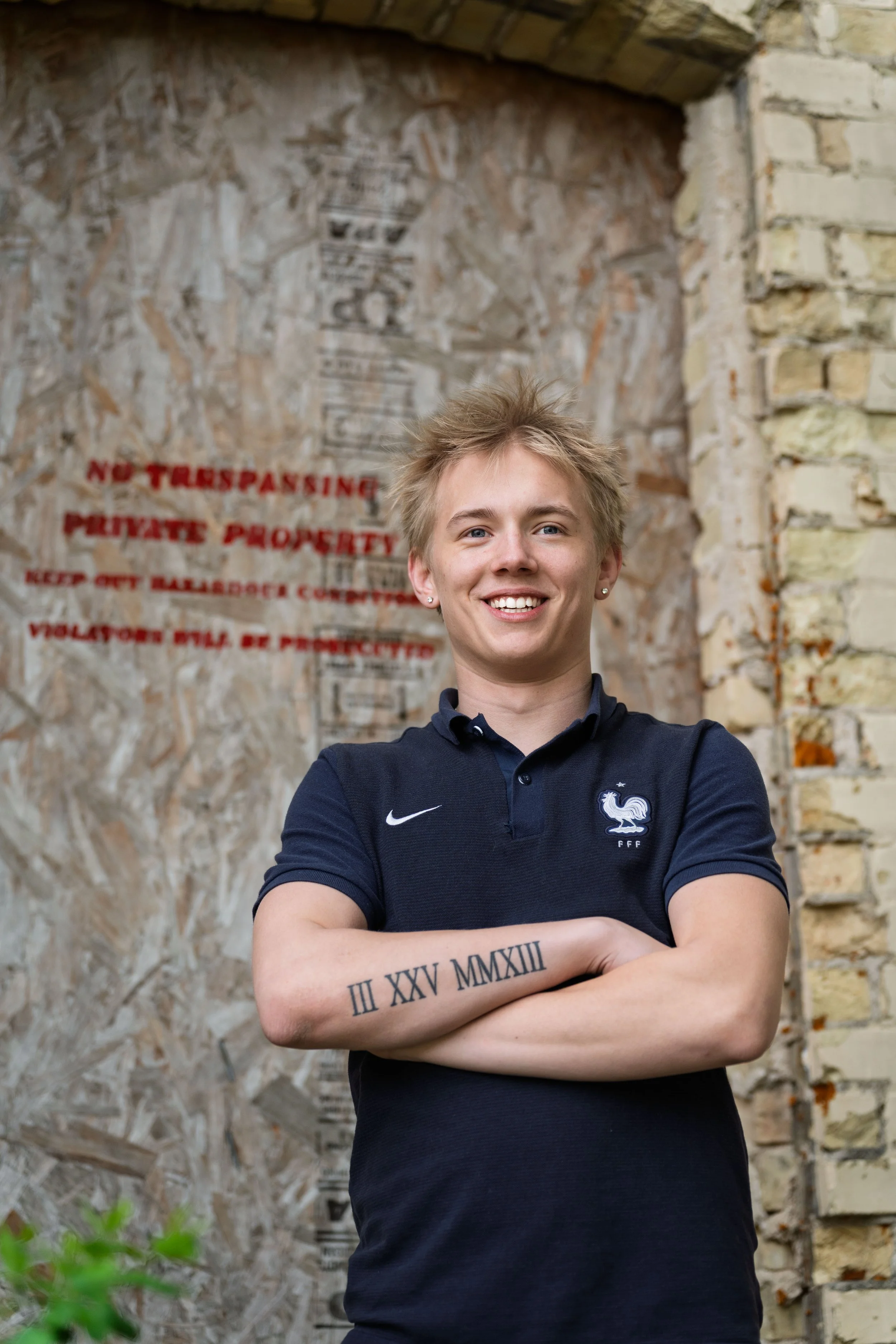 A young person with short blond hair, wearing a navy blue polo shirt with a French national football team emblem, confidently smiling with arms crossed in front of a brick wall with a 'No Trespassing' sign.