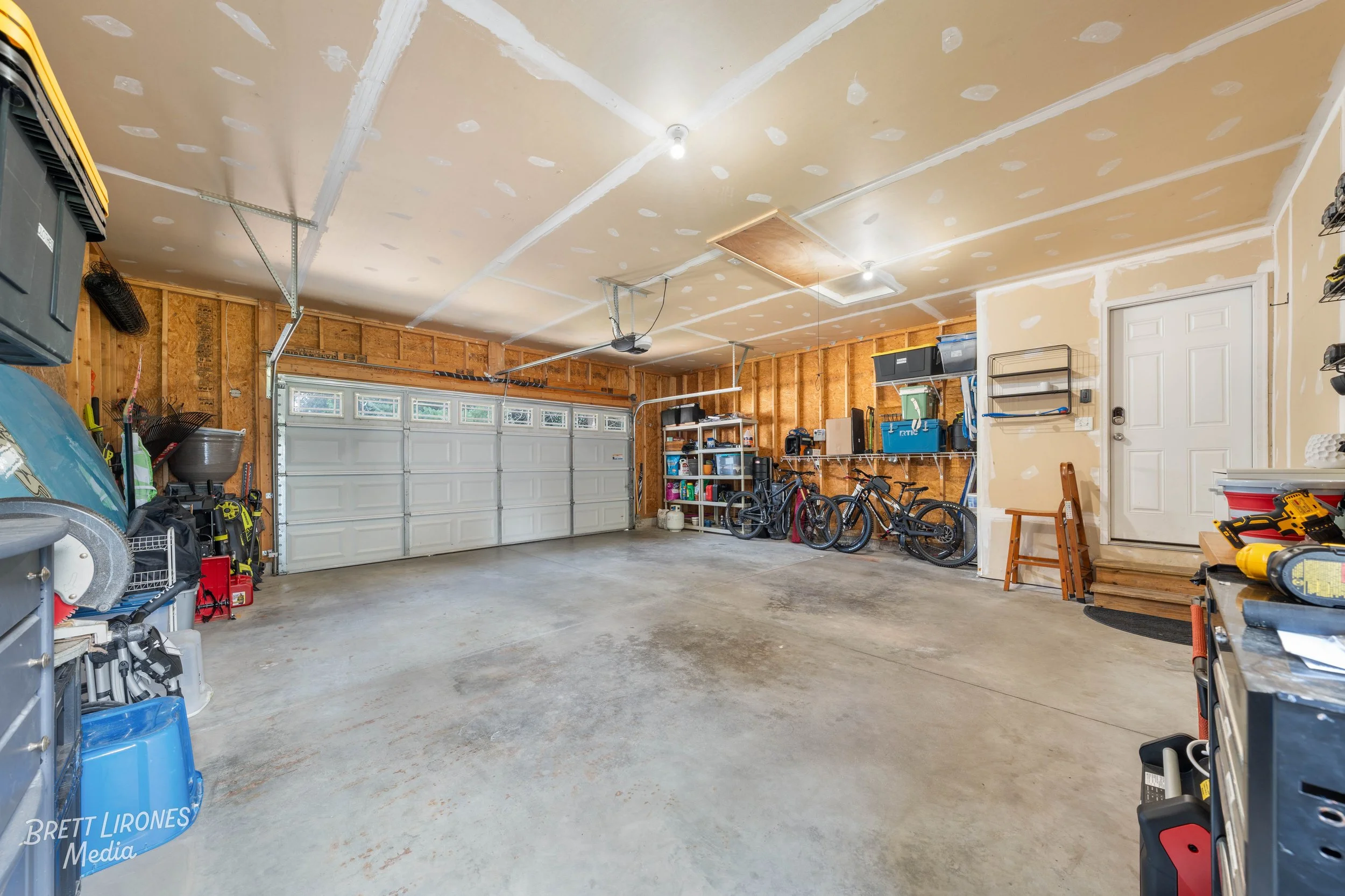 An empty garage with a concrete floor, unfinished drywall ceiling, and organized storage shelves along the back wall. Bicycles, storage boxes, and tools are stored on the right and left sides.
