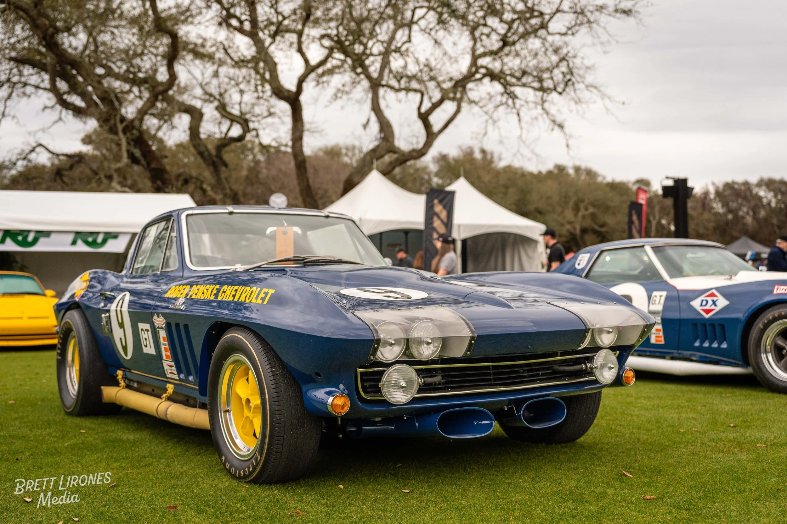 A vintage blue race car with yellow accents, number 9, at an outdoor car show. There are tents and other classic race cars in the background.