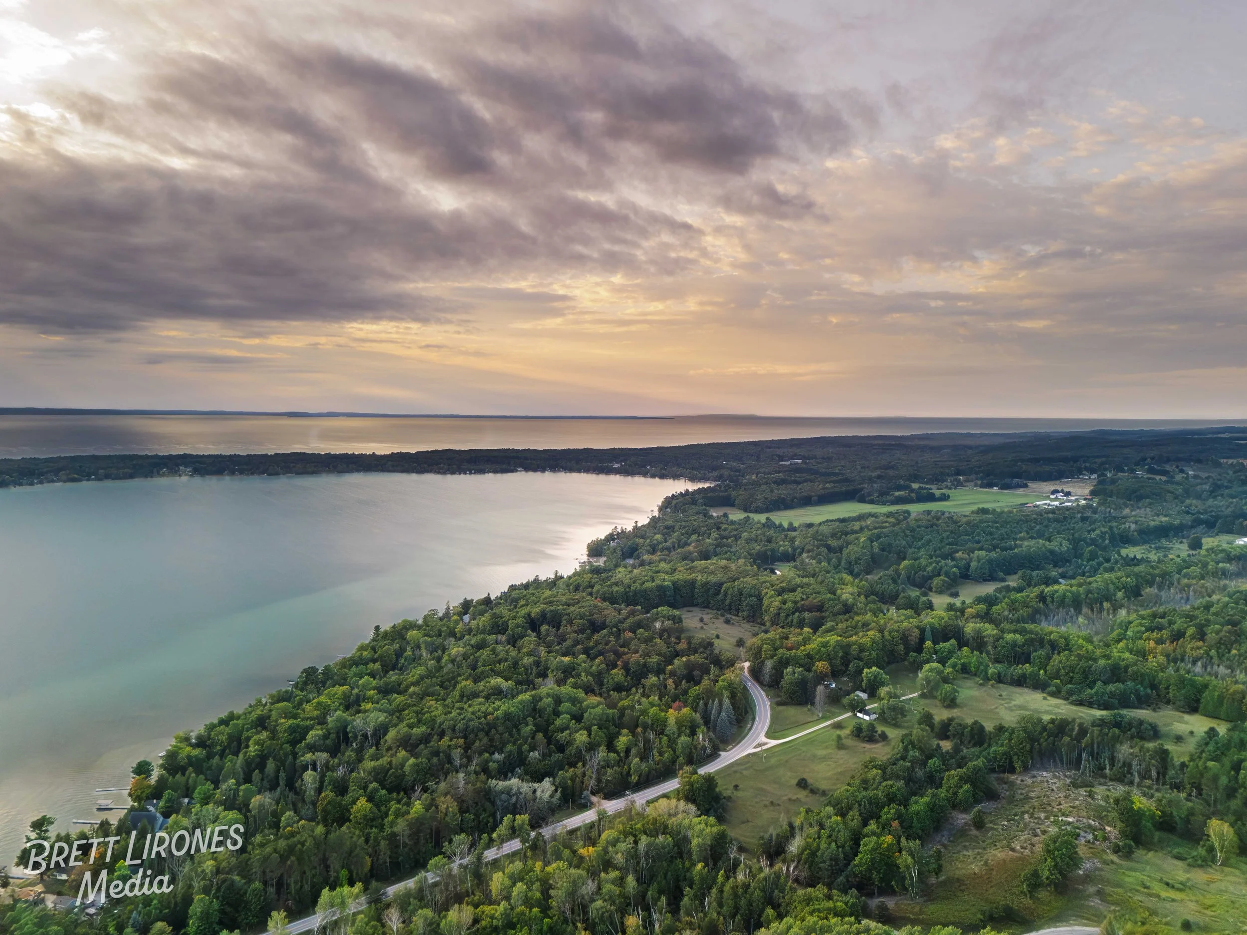 Aerial view of a lake and surrounding lush green forests and fields under a cloudy sky during sunset.