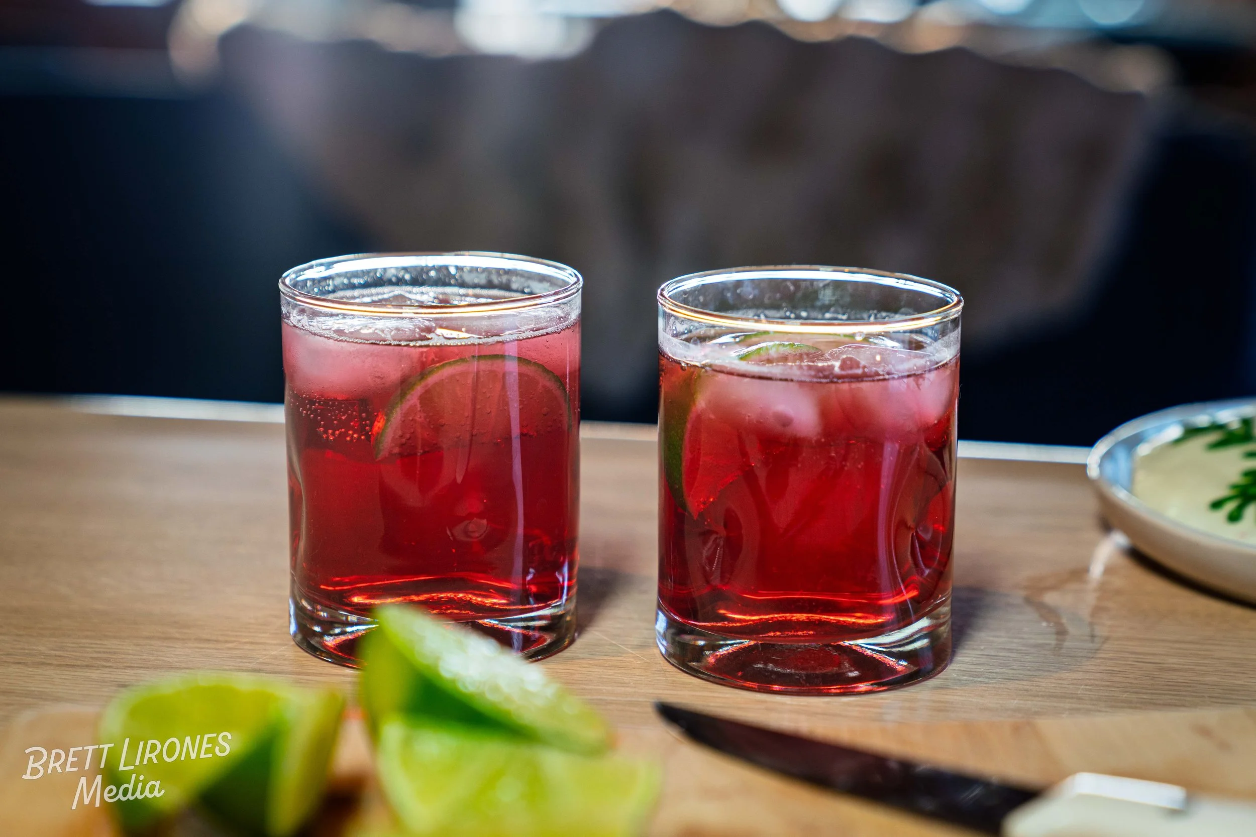 Two glasses of red-colored beverage garnished with lime slices on a wooden table with lime wedges and a plate in the background.
