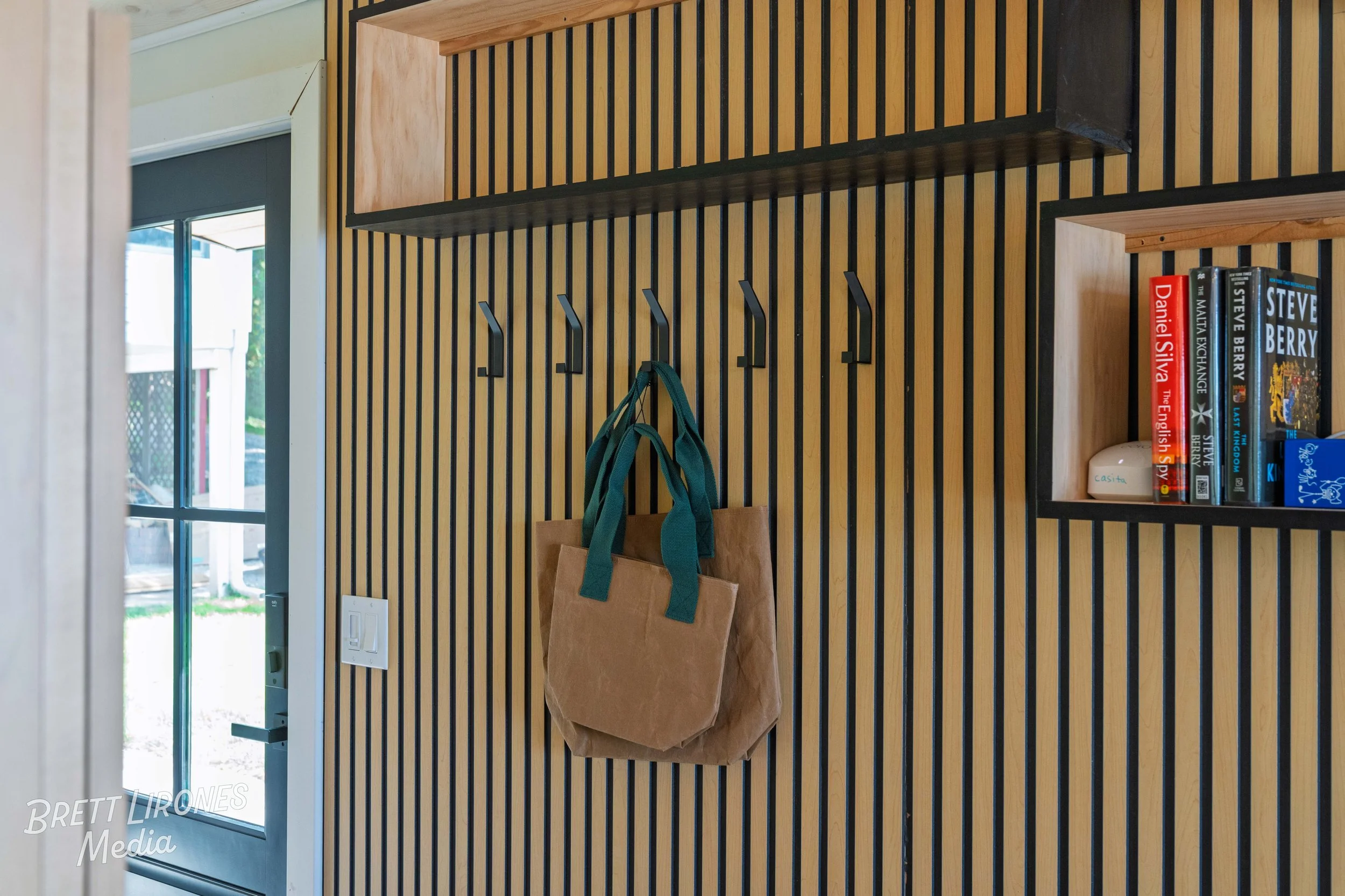 Wooden wall with black vertical slats, hooks with bags hanging, and a small shelf with books and a white object. Part of a door and window are visible.
