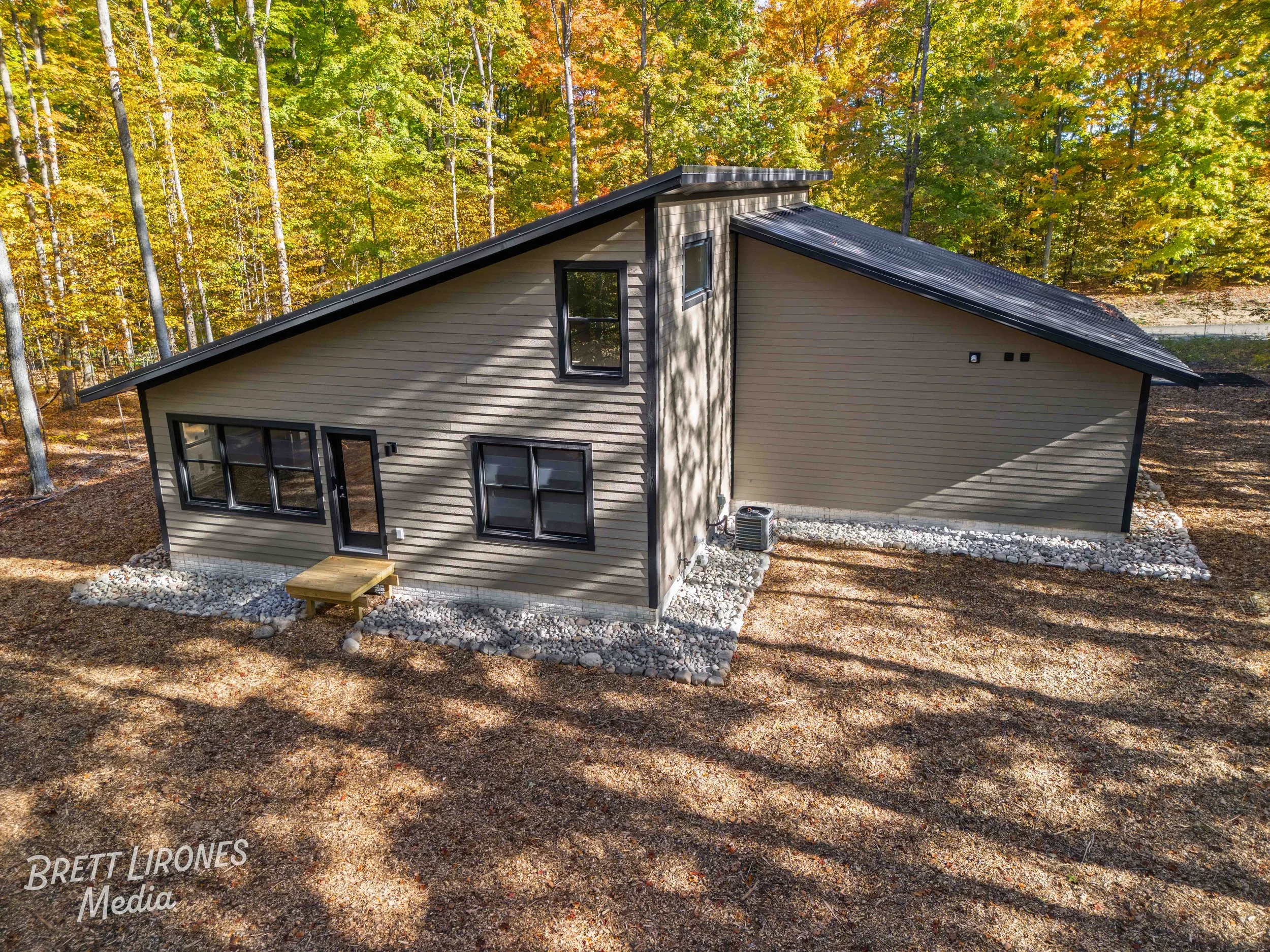 Modern house with tan siding and black trimmed windows in a wooded area with fall foliage.