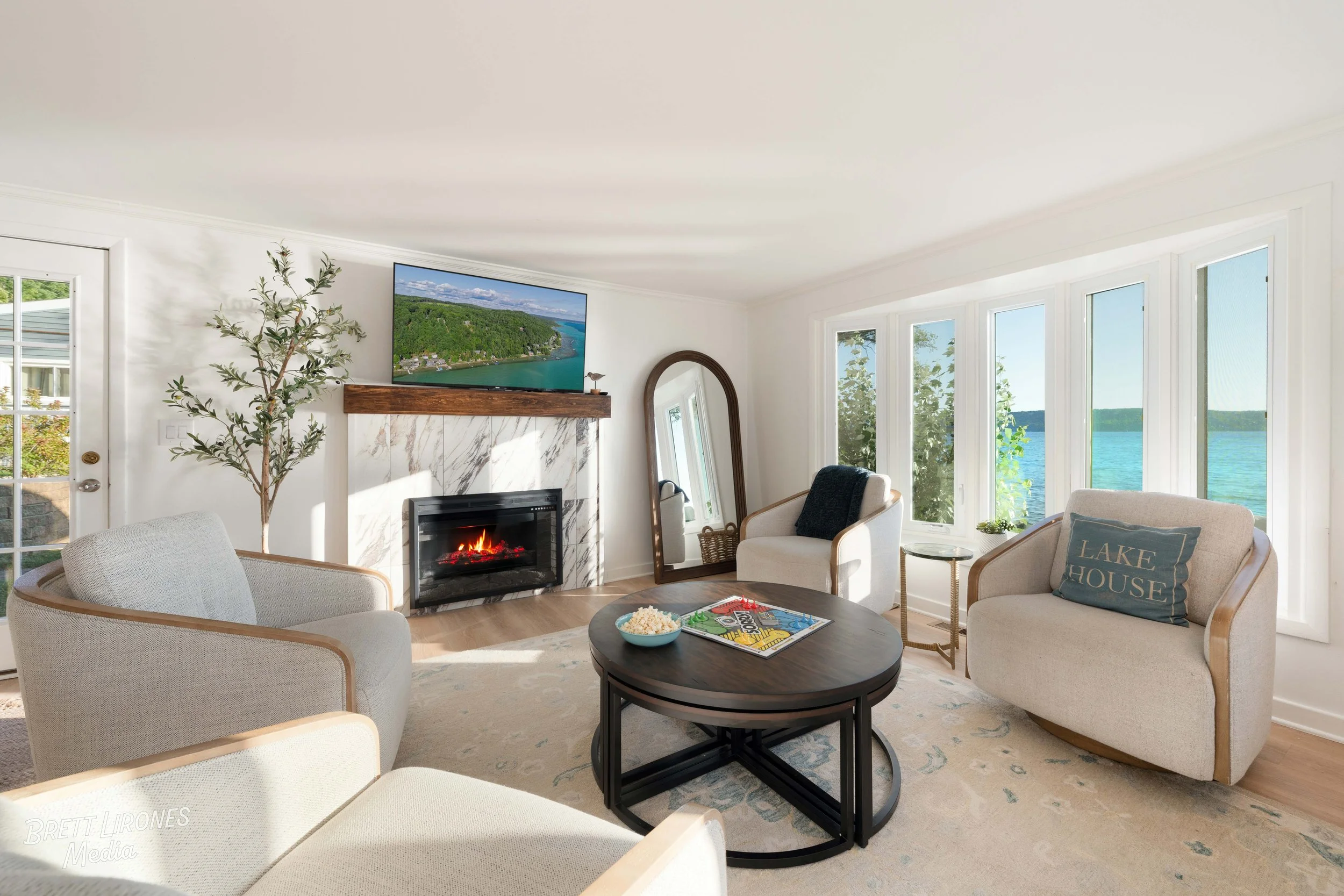 Living room with ocean view, white walls, fireplace, flat-screen TV, armchairs, coffee table with magazines and popcorn, and large windows overlooking the lake.